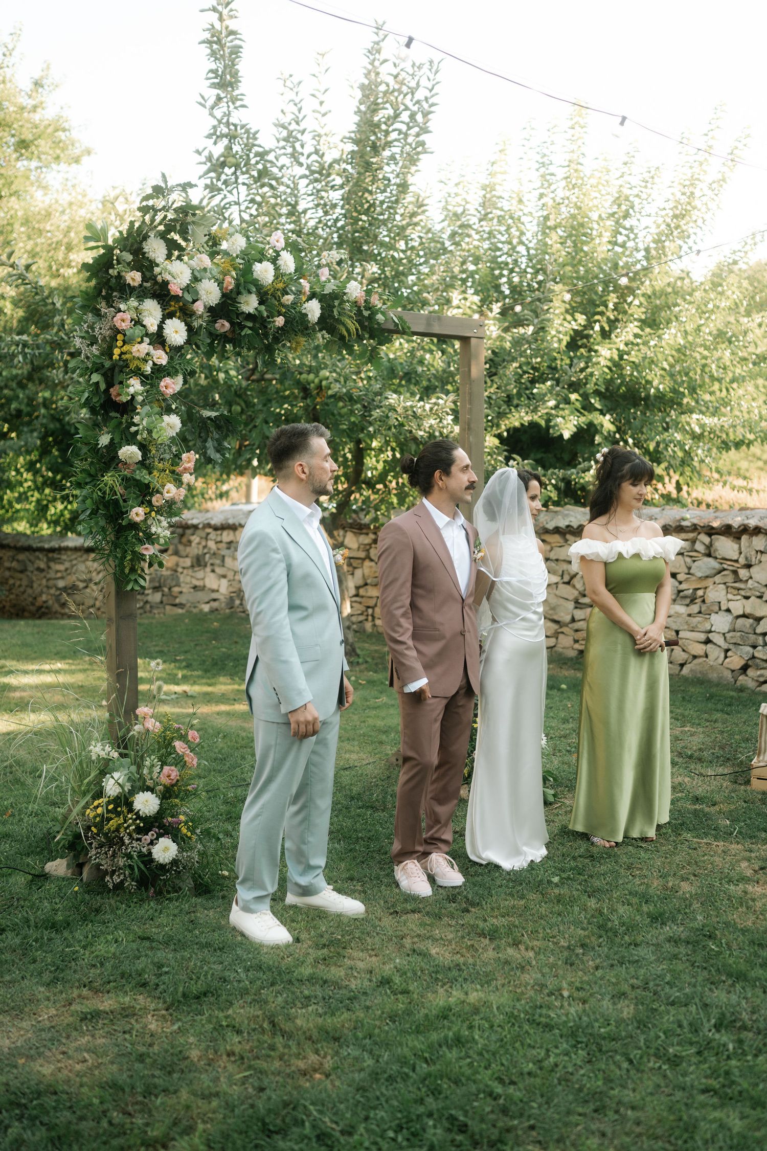 Wedding party members in pastel and neutral tones pose together in a garden setting with floral arrangements.