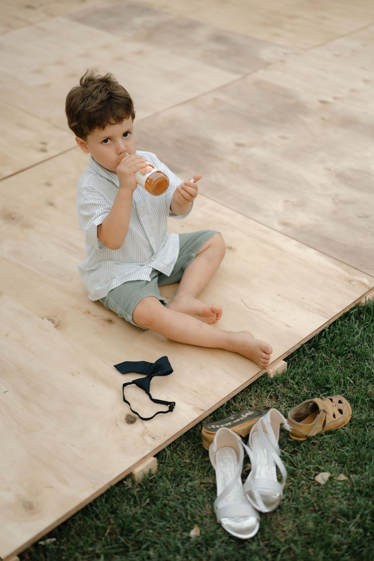 A young child sits on a wooden platform next to grass with shoes and socks scattered nearby.