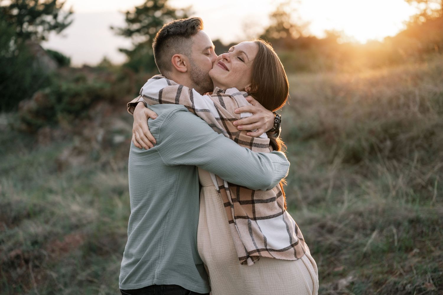 A couple shares a romantic embrace and kiss at sunset in a natural outdoor setting with golden light.