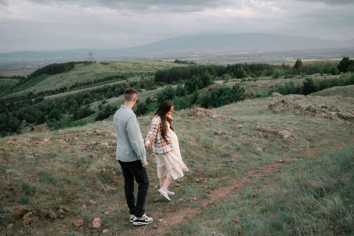Couple walking along a scenic mountain trail overlooking rolling hills and valleys on an overcast day.