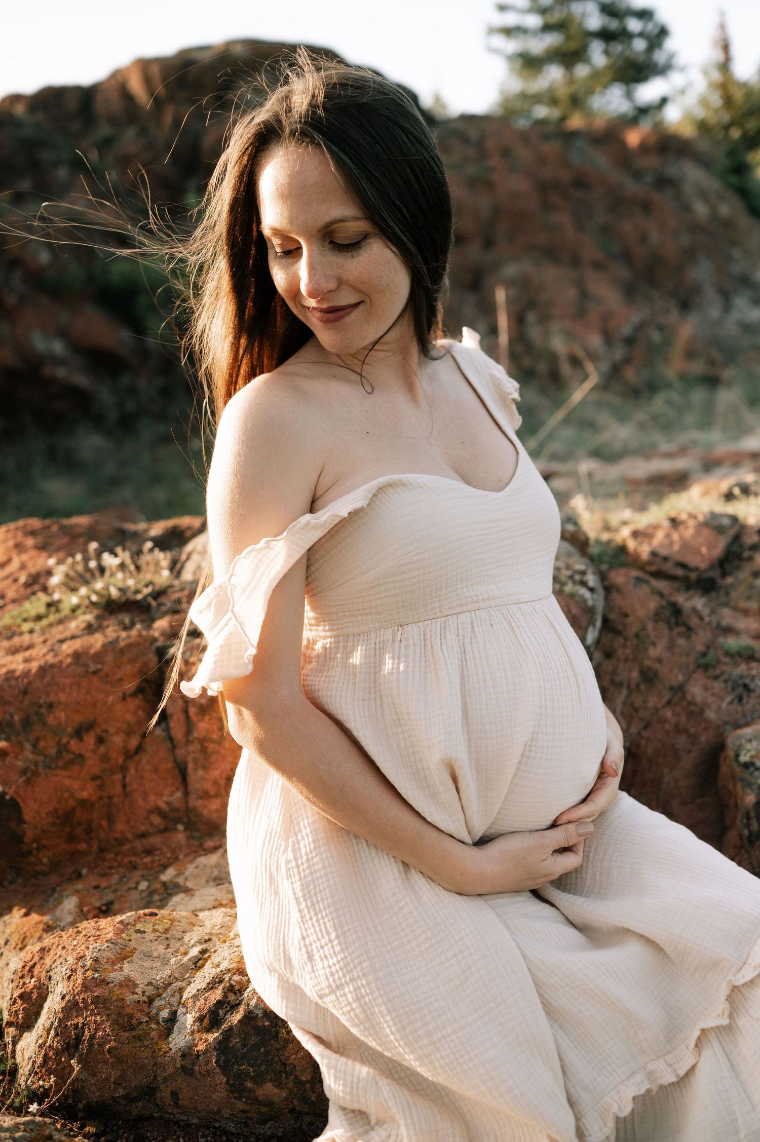 A woman in a flowing white dress sits gracefully on red rocks during golden hour in a natural outdoor setting.