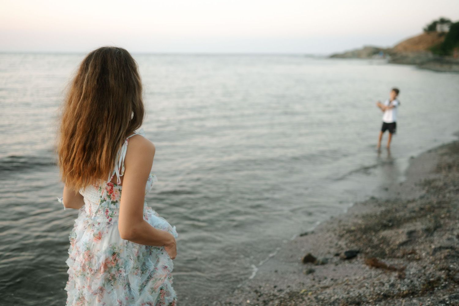 A couple walks along a misty beach shoreline at sunset holding hands while waves gently lap at their feet.