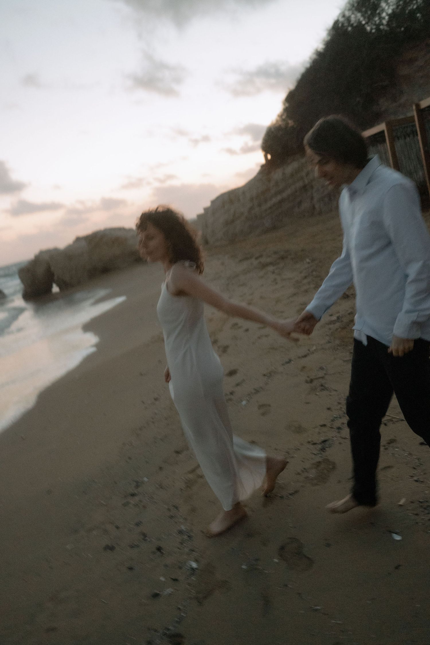 Couple holding hands walking barefoot on a sandy beach at dusk, woman in white dress, cliffs in background.