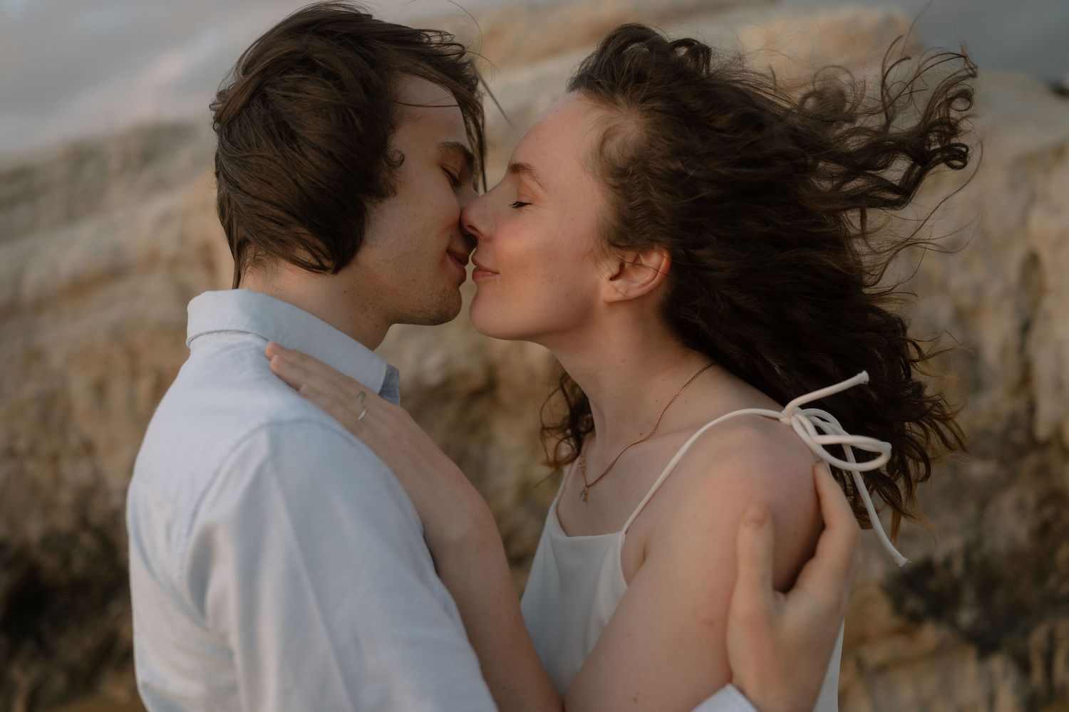 Romantic couple sharing a kiss outdoors with wind-blown hair against a scenic rocky backdrop.