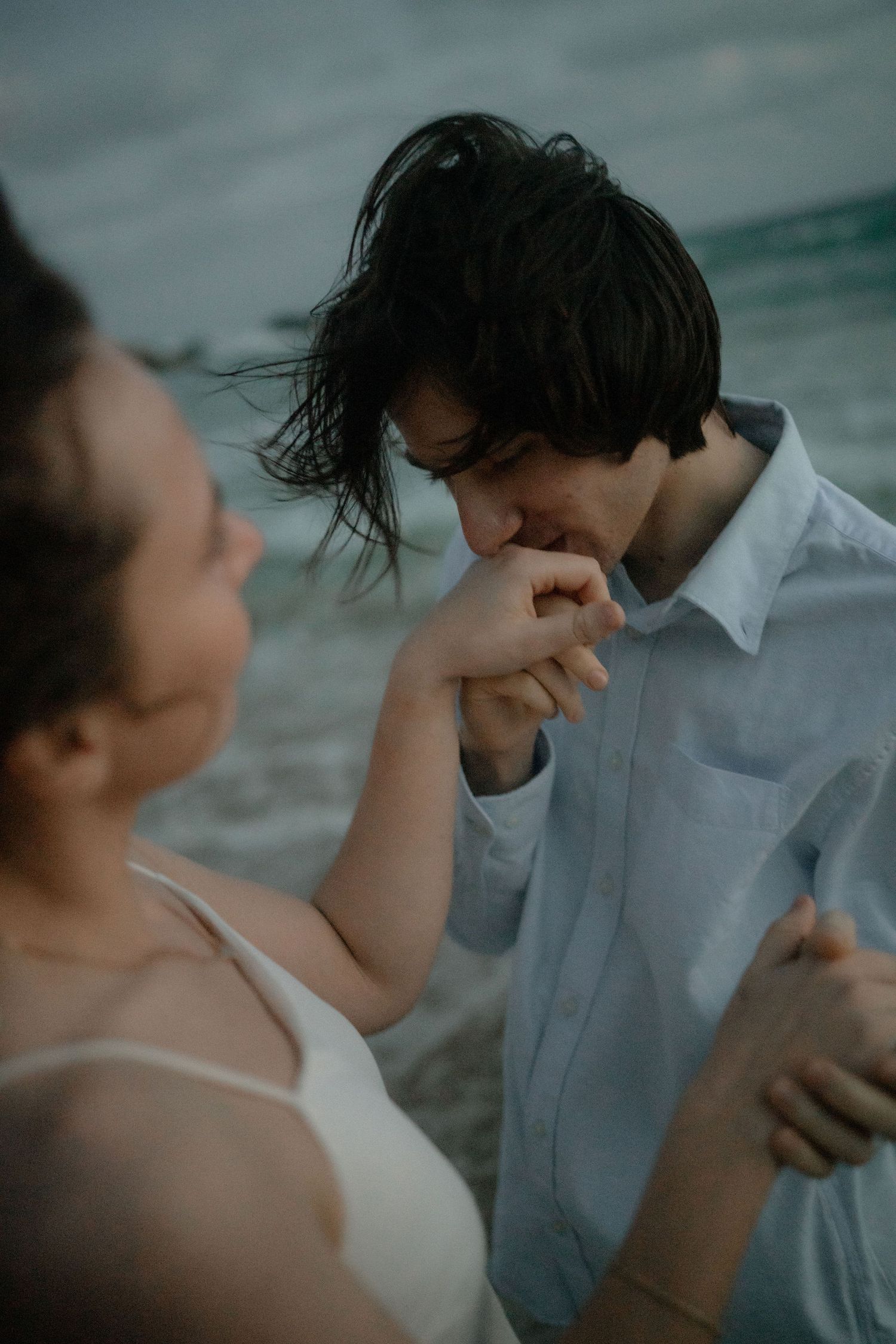 A man in a blue shirt kissing a woman's hand on a beach at dusk.