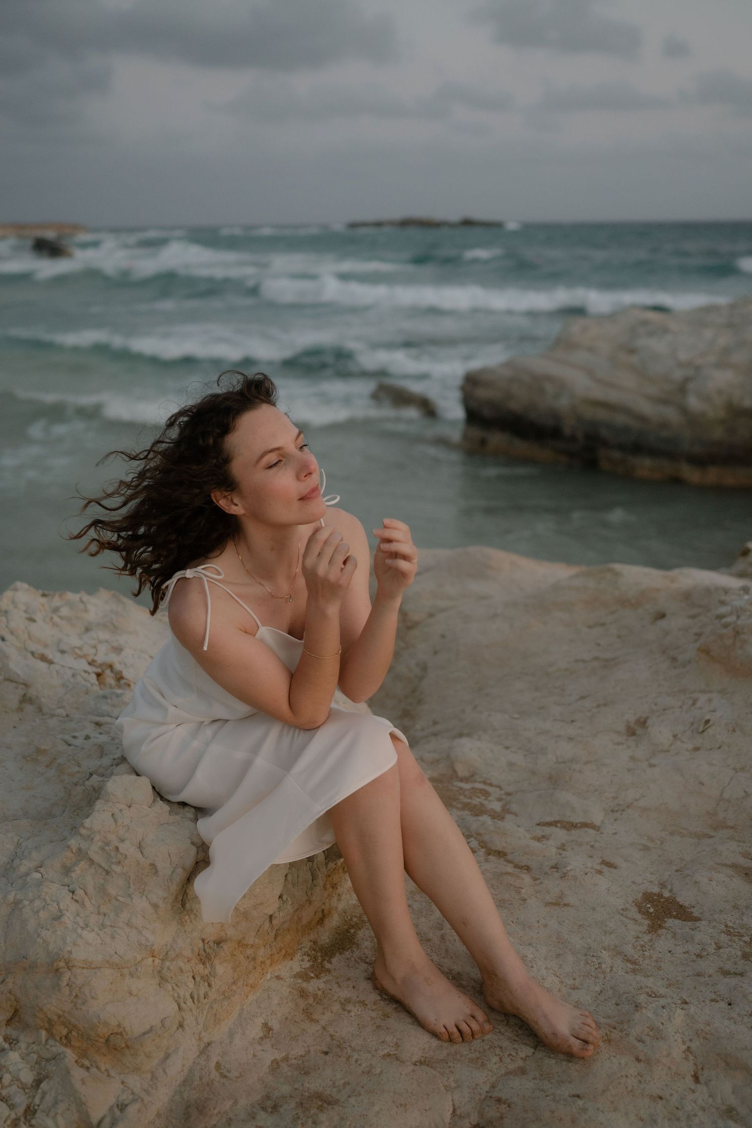 A barefoot woman in a white dress sits on rocky shore, hair blowing in wind, with turquoise ocean waves behind her.