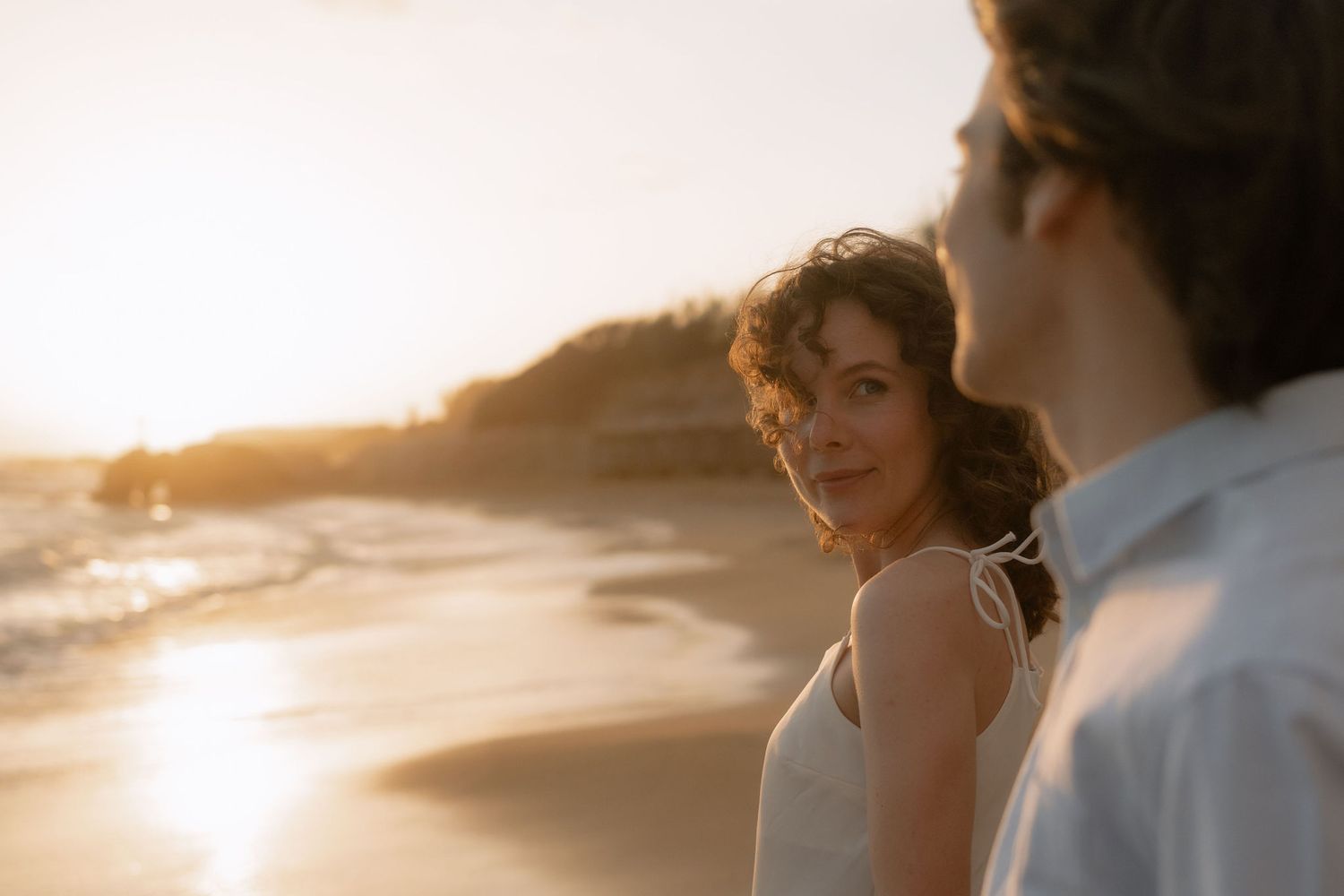 Woman in white dress smiling at sunset on a beach with golden hour light and ocean waves in background.