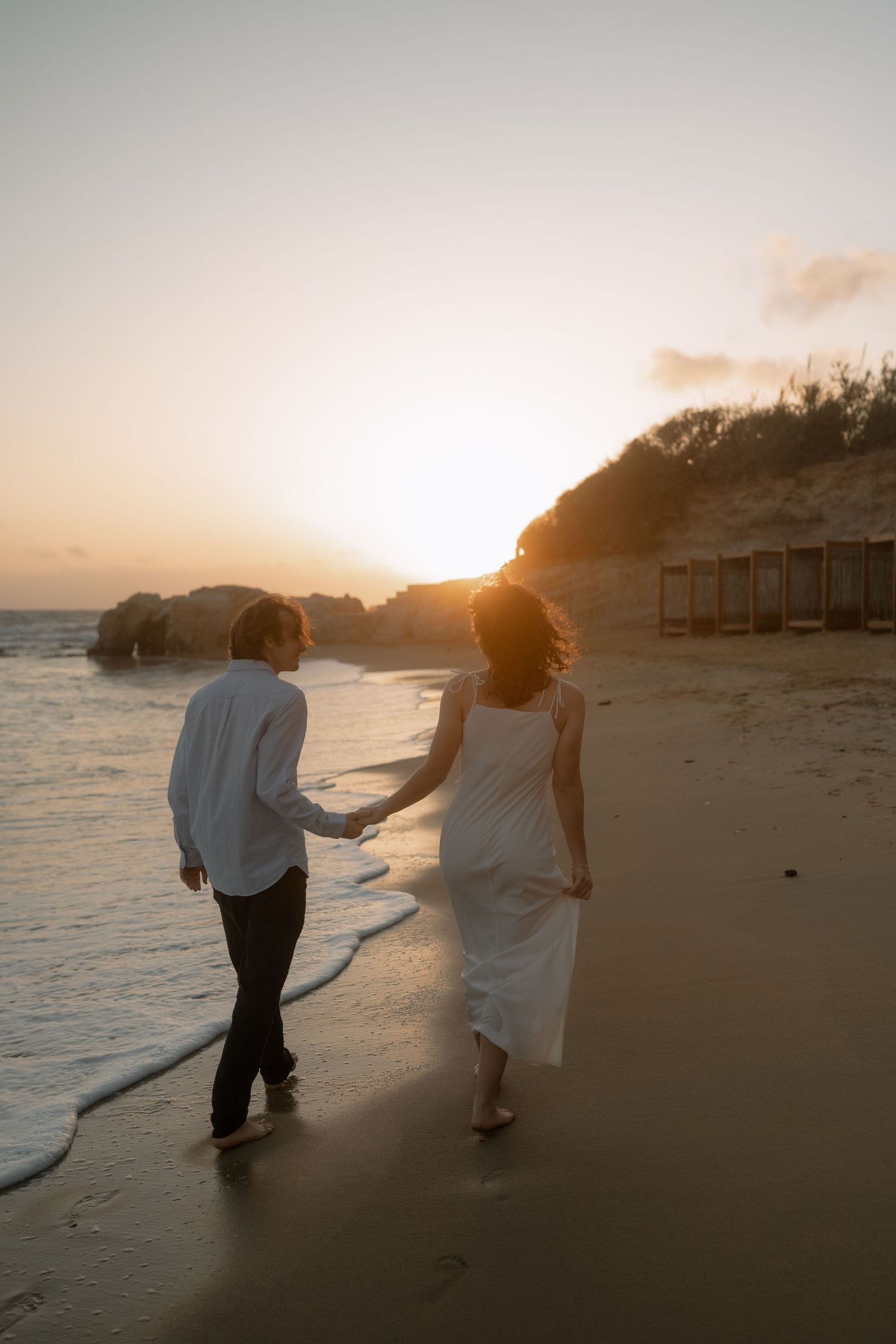 Couple holding hands walking on a sandy beach at golden sunset, woman in white dress, romantic seaside stroll.