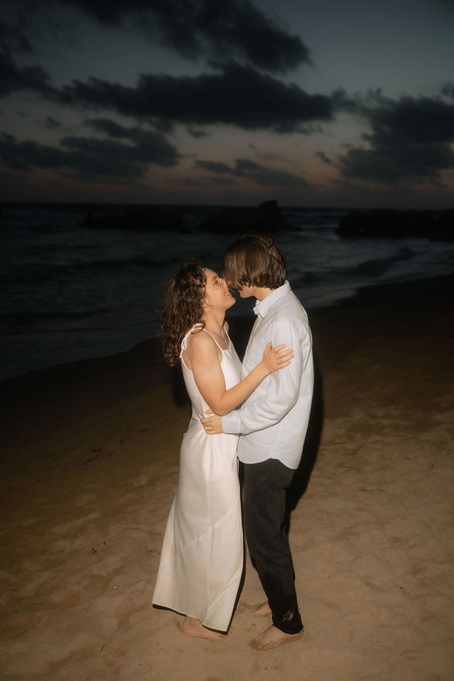 Couple sharing a romantic moment on a sandy beach at dusk, woman in white dress embracing man near ocean waves.