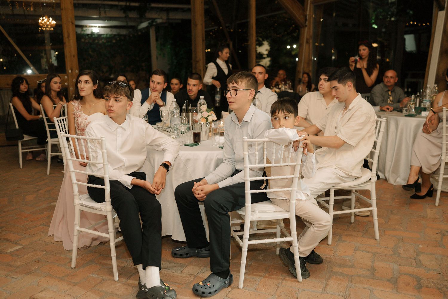 Wedding guests seated at elegant white tables during an indoor reception share laughter and conversation.
