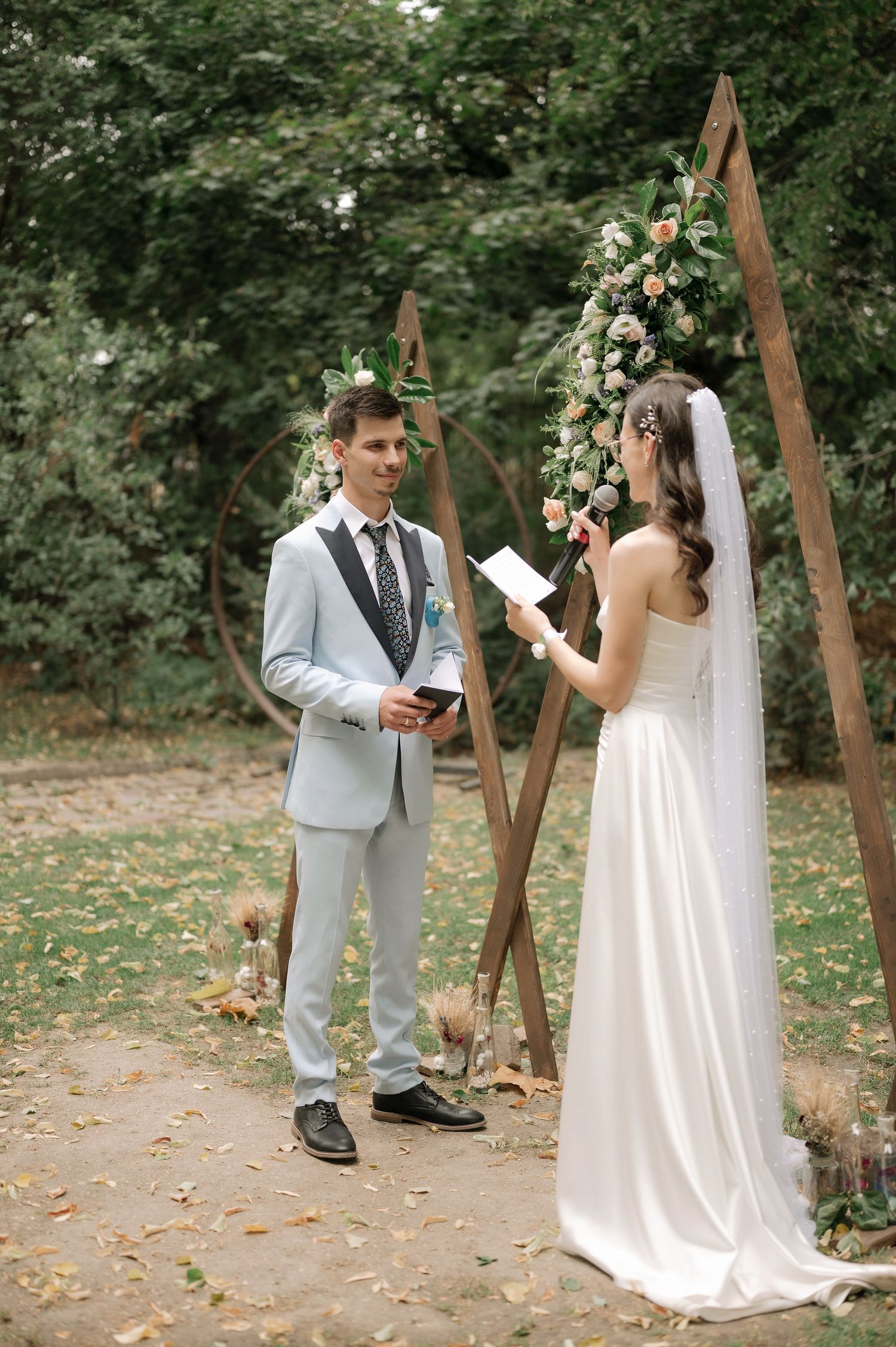 A couple exchanges vows during an outdoor wedding ceremony under a wooden arch decorated with white flowers.