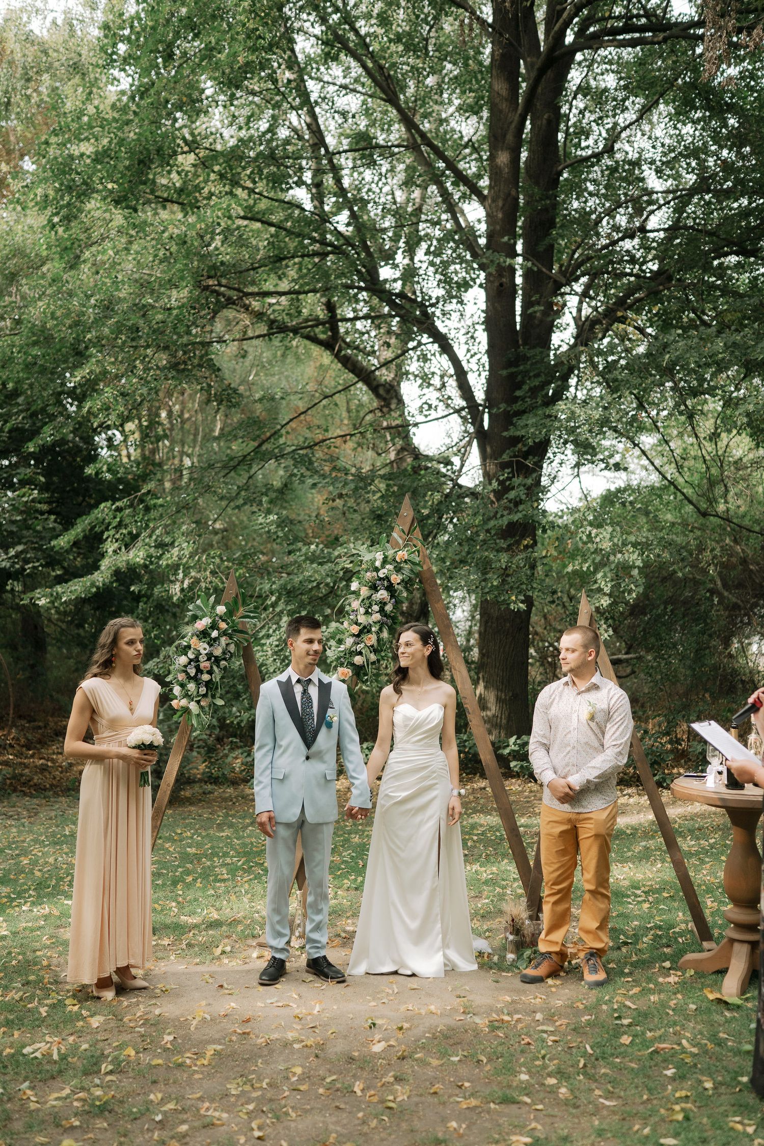 Outdoor wedding ceremony in a wooded setting with guests gathered among autumn leaves on the ground.