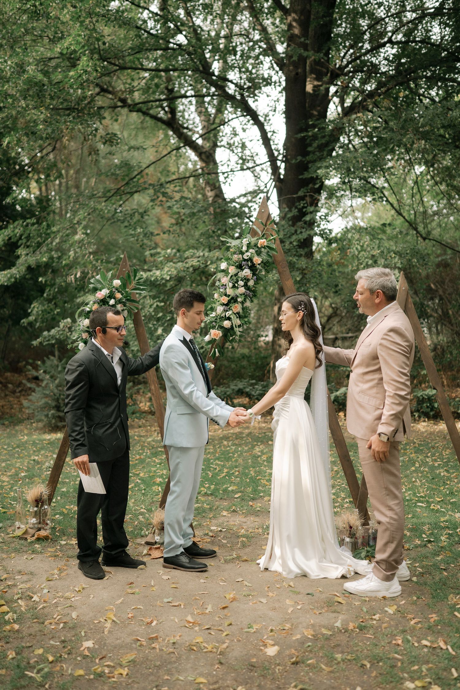 An intimate outdoor wedding ceremony takes place in a wooded setting with autumn leaves scattered on the ground.