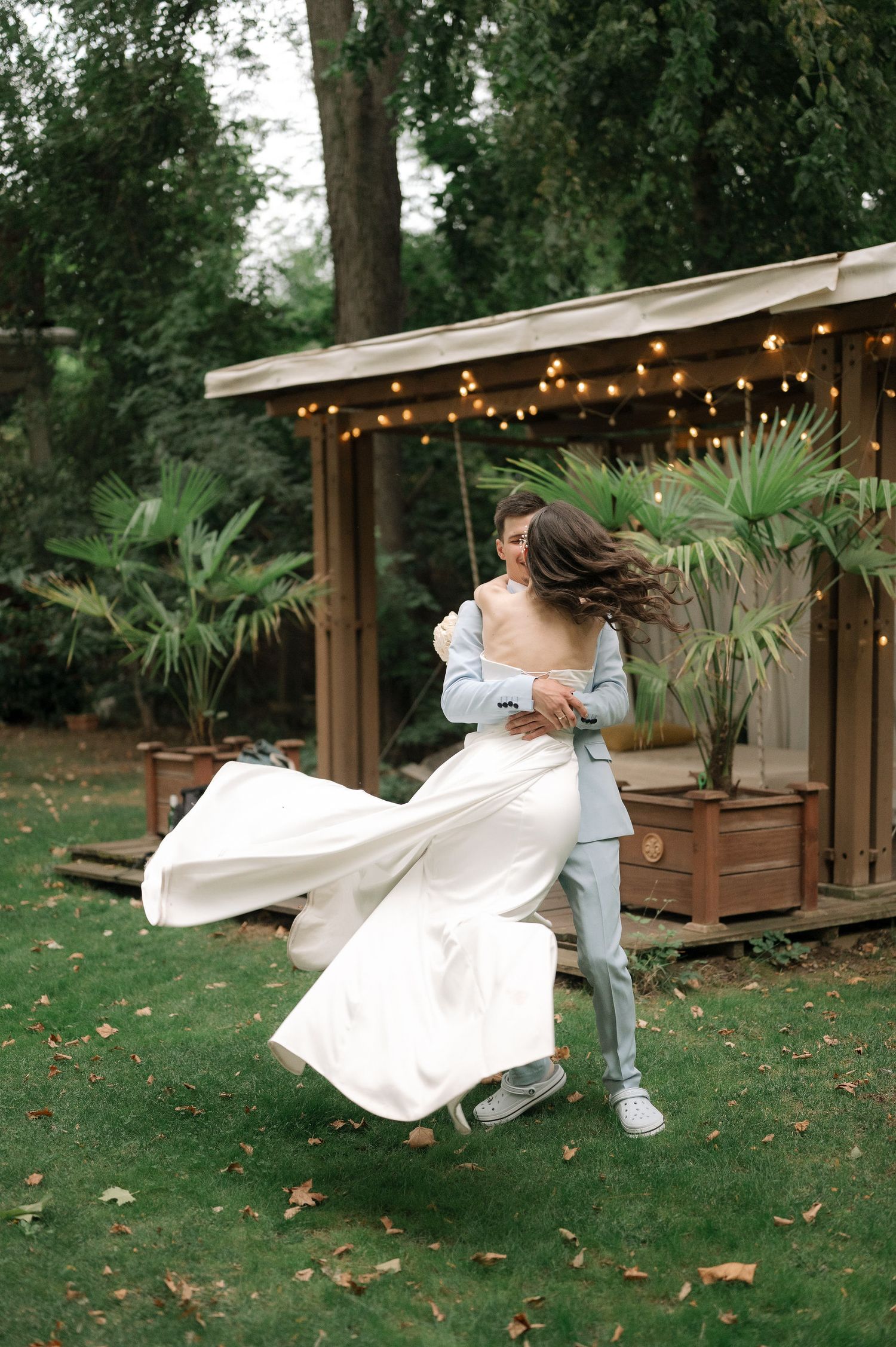 A couple performs a playful dance lift in a garden setting with string lights and palm plants in the background.