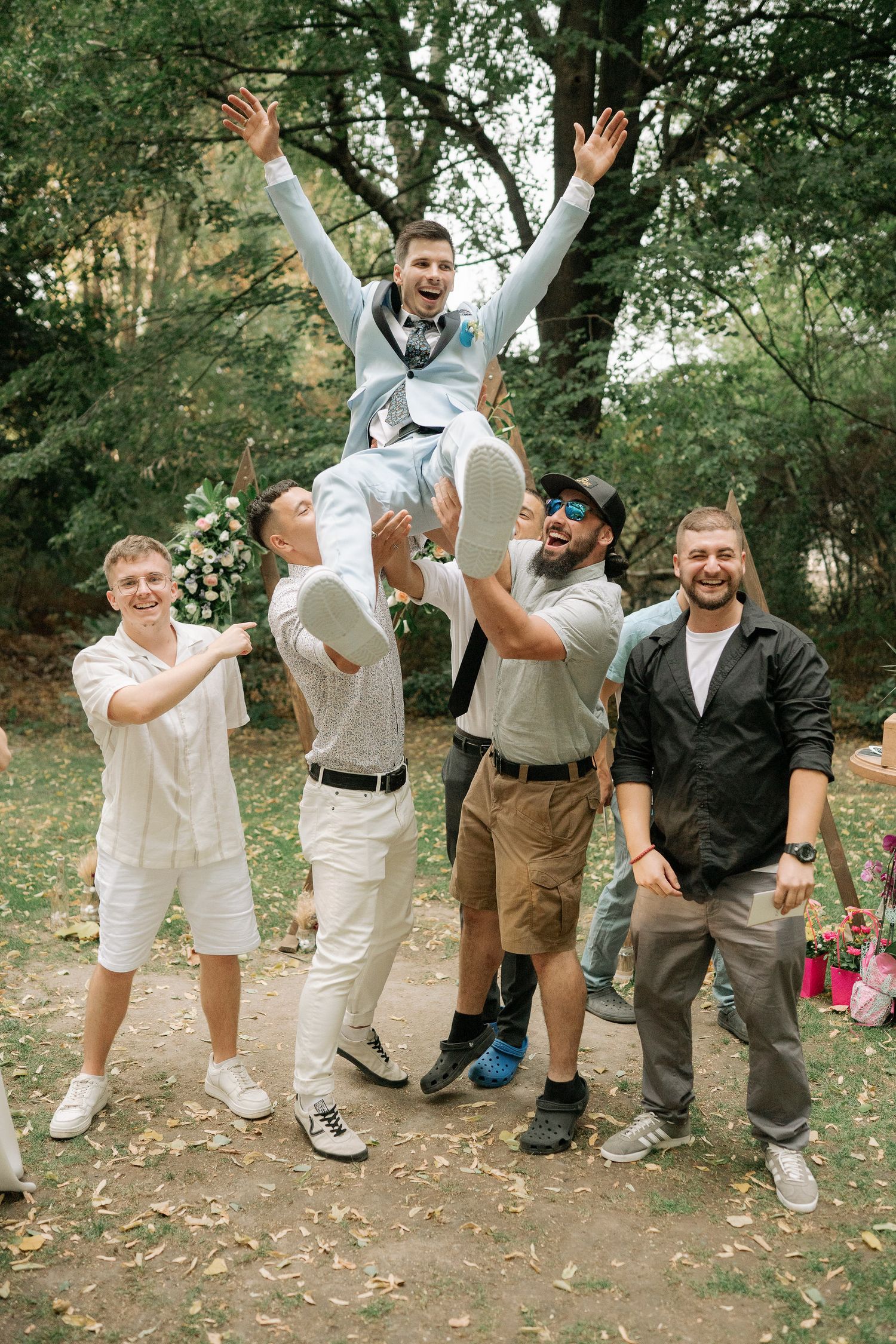 A group of people in coordinated white and brown outfits jump joyfully in a wooded outdoor setting during a celebration.