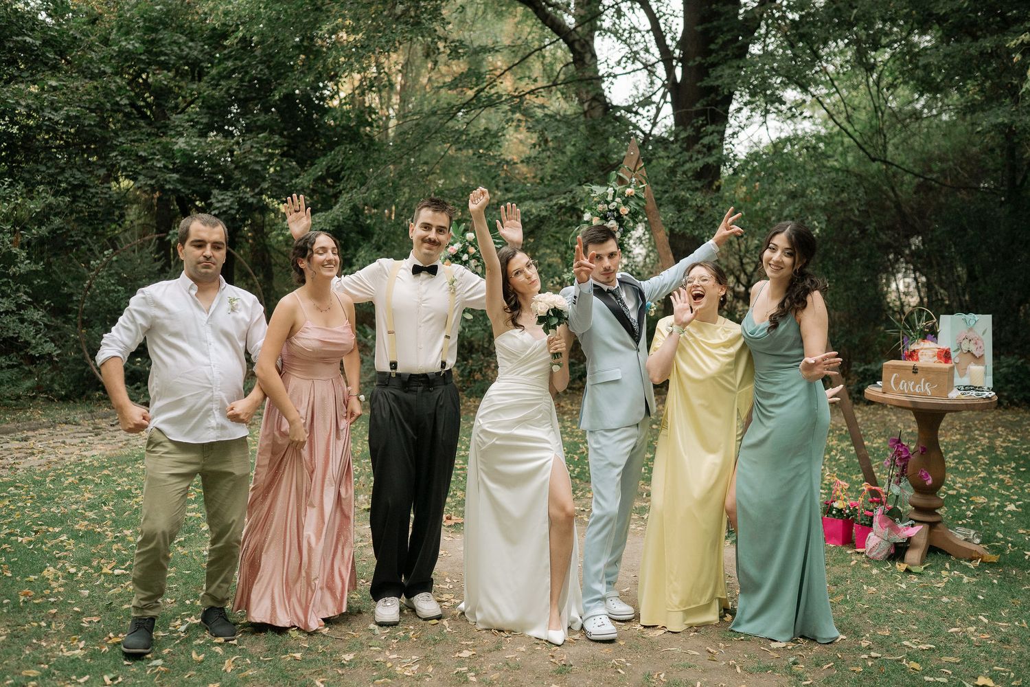 Wedding party in pastel dresses and formal attire celebrate joyfully in an outdoor garden setting.