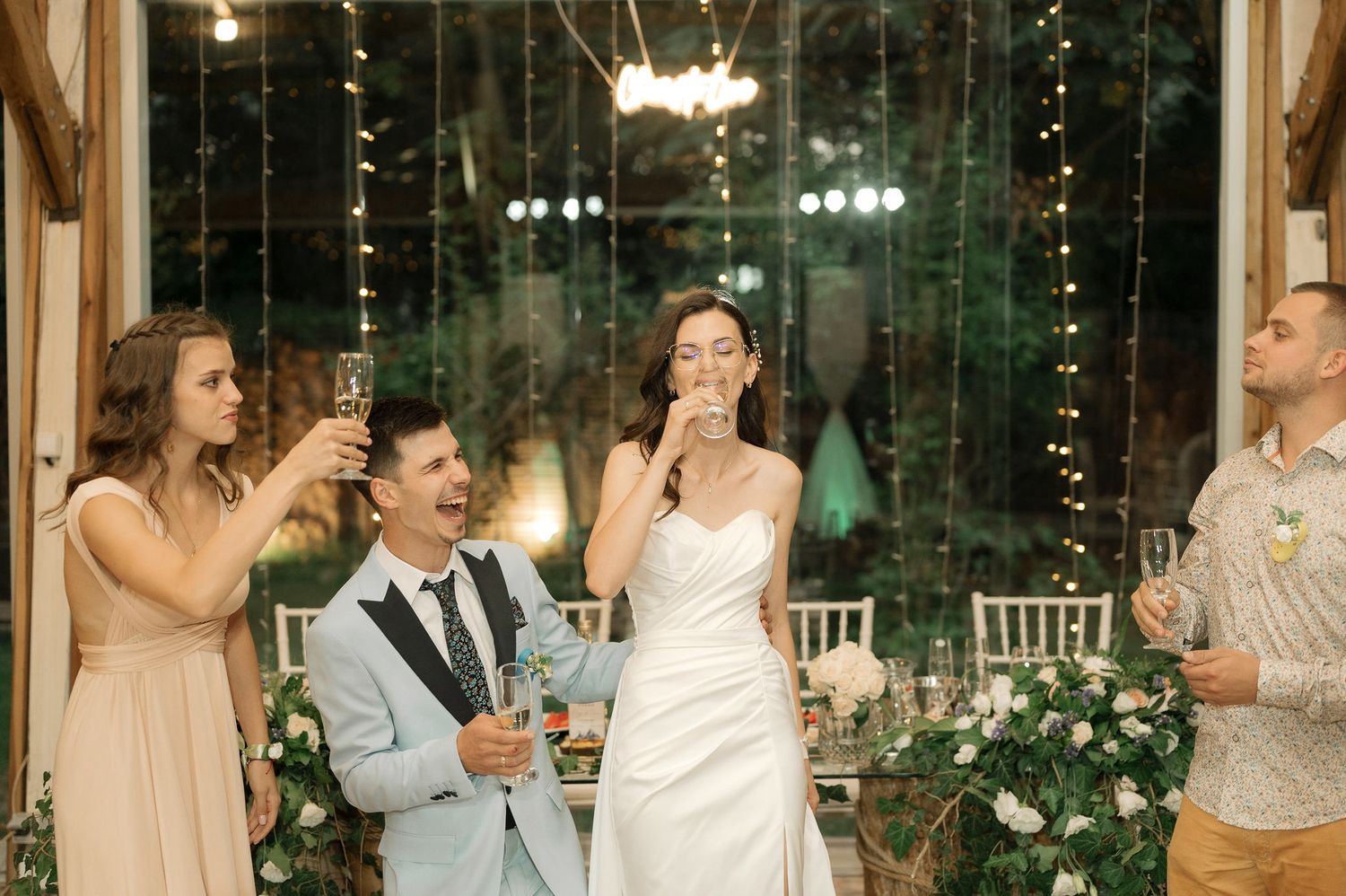 Wedding party laughs together during reception celebration with twinkle lights and greenery decorations in background.