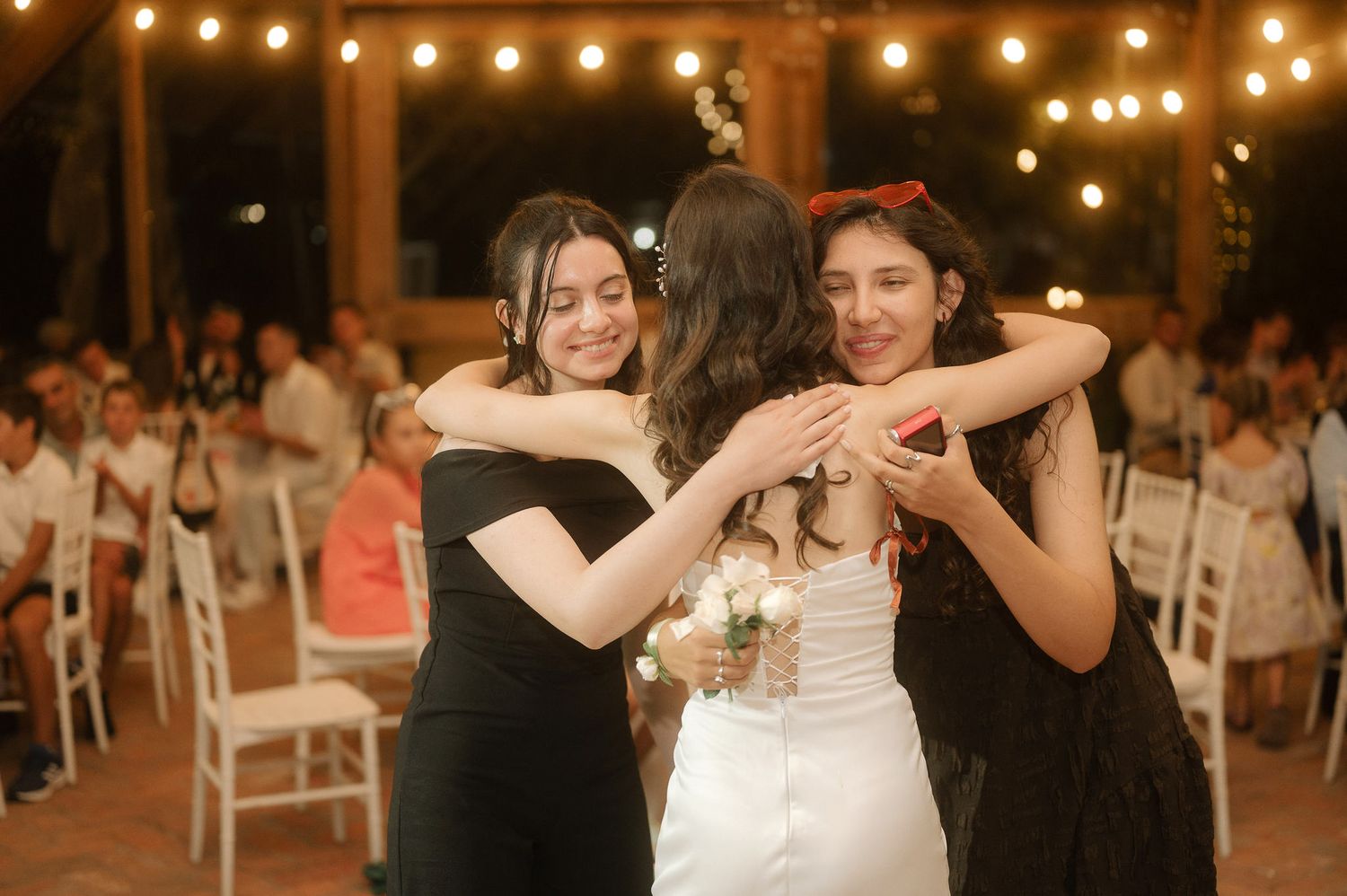 Three people share an emotional embrace on the dance floor at a wedding reception under warm lighting.