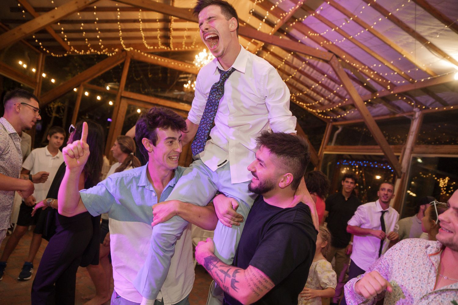 Wedding guests celebrate and dance together at a rustic barn reception venue illuminated with string lights.