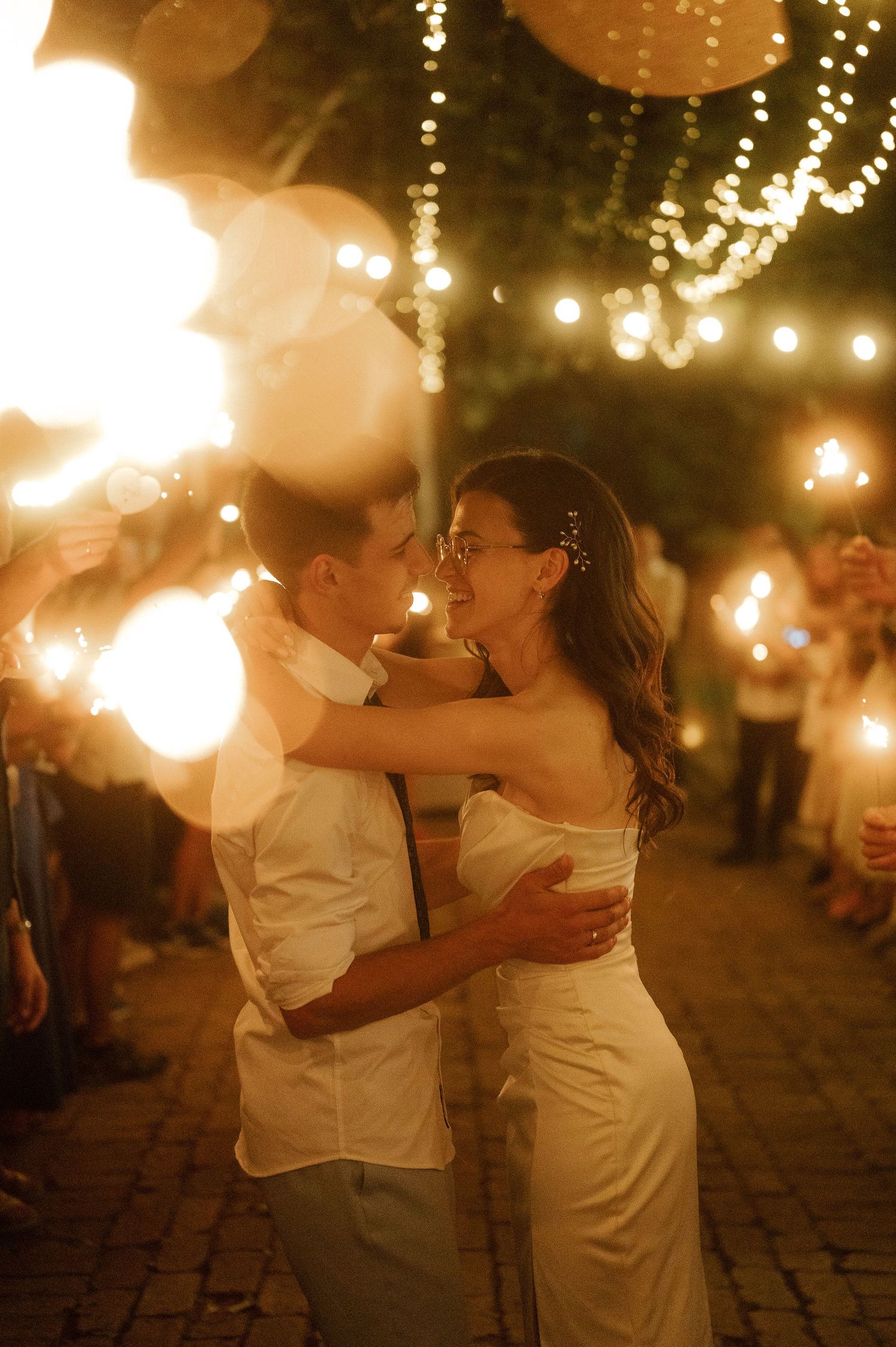 A romantic couple embraces and dances under warm glowing string lights on a cobblestone patio at night.