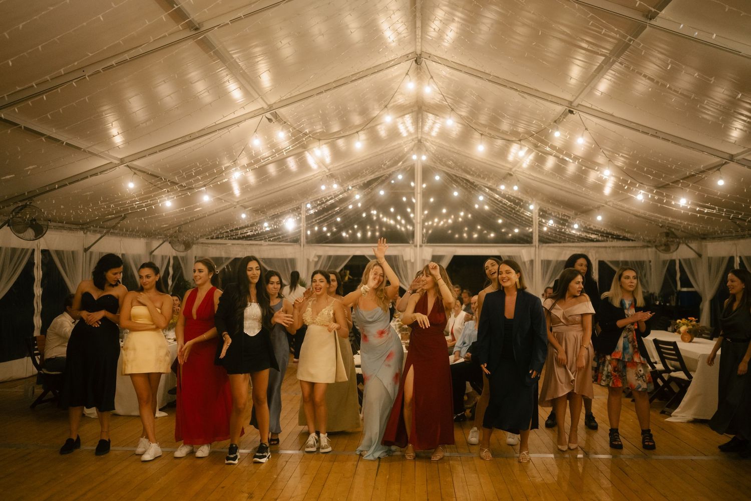 Panoramic view of a wedding reception tent with string lights illuminating guests dancing on the wooden floor.