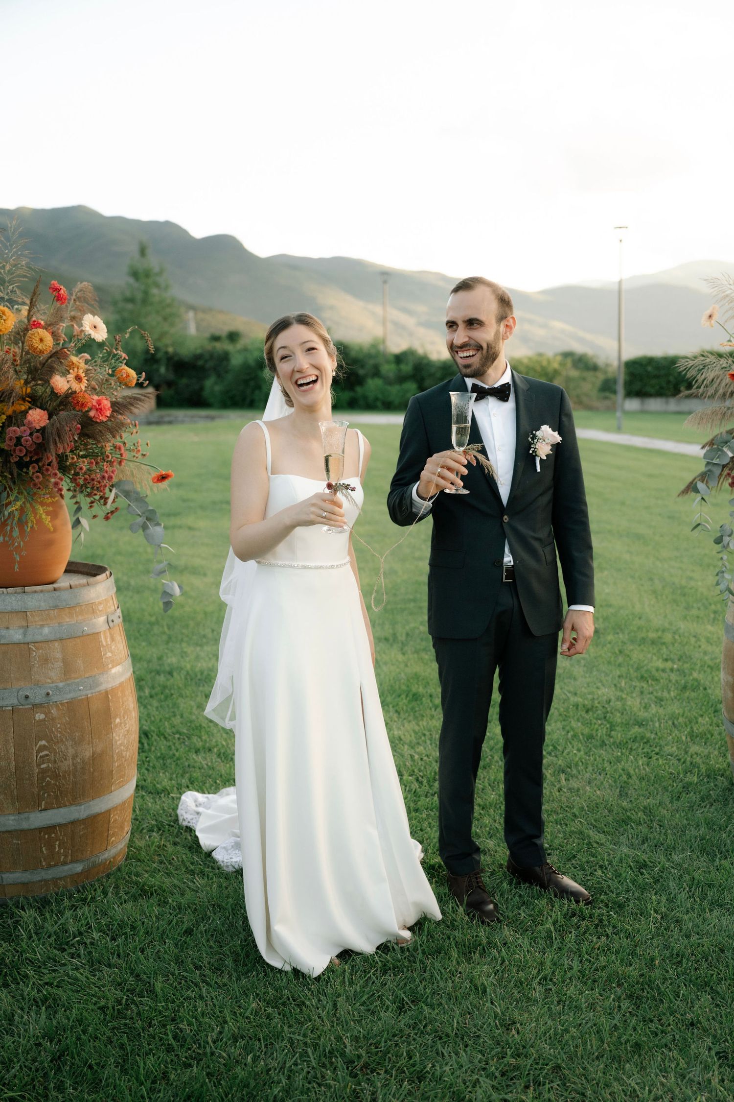 A couple in wedding attire shares a laugh while holding wine glasses on a scenic grassy hillside with wine barrels.
