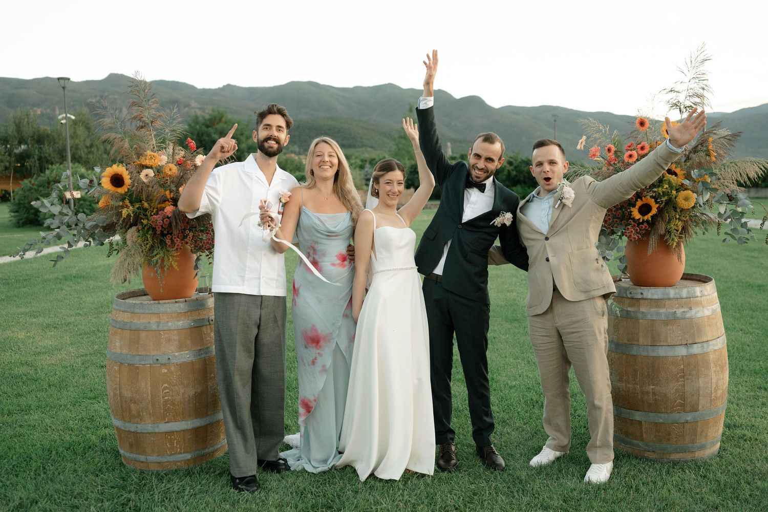 Wedding party celebrates outdoors with rustic wine barrels and sunflower arrangements on a green lawn with mountain backdrop.