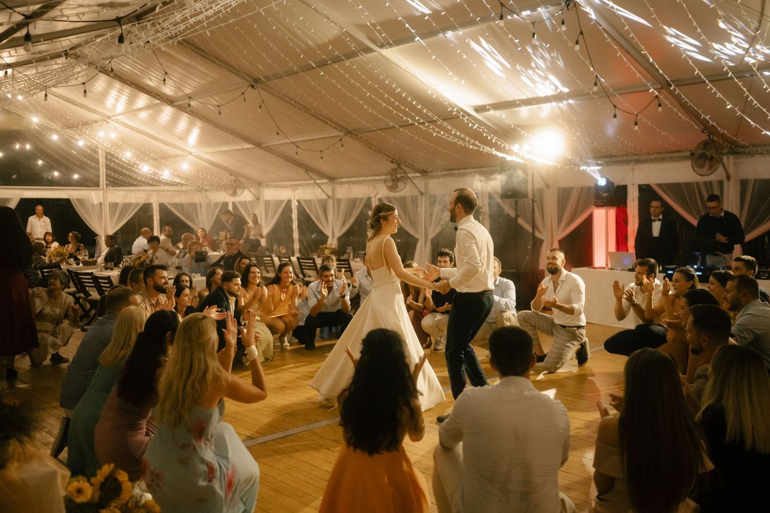 A crowded dance floor filled with people dancing and celebrating at an indoor reception venue with warm lighting.