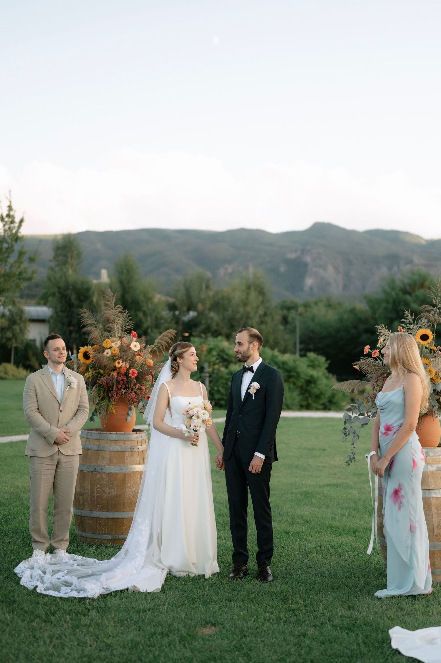 Wedding party gathers on green lawn with rustic wine barrel and mountain backdrop at outdoor ceremony venue.