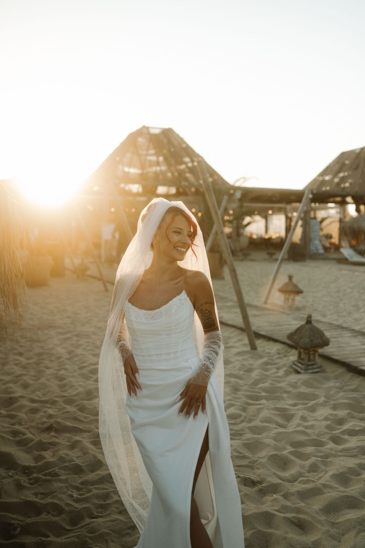 A bride in an elegant white dress with a flowing veil stands on a beach with rustic structures in the background.
