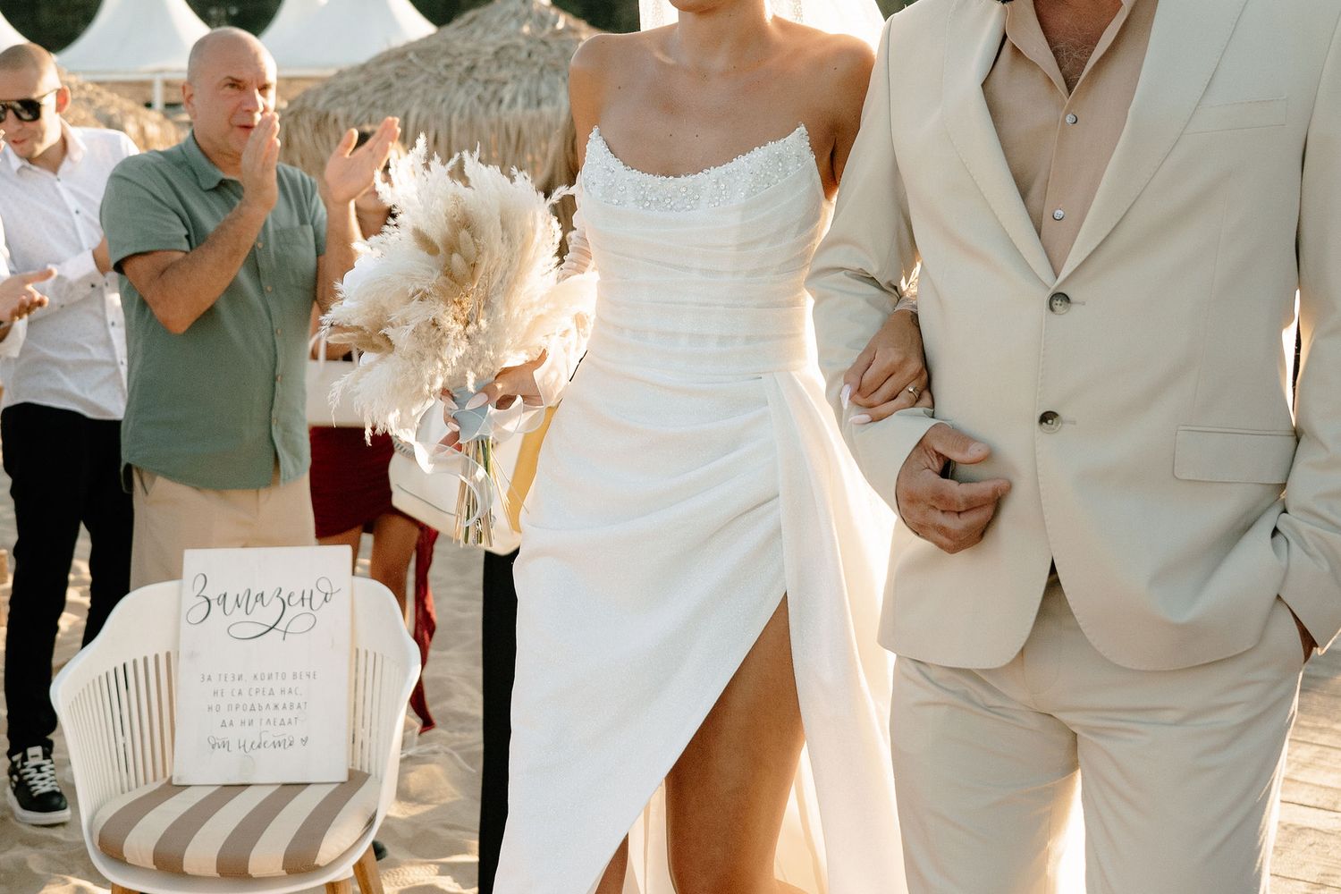 Wedding party members in white and beige attire walk together on a coastal boardwalk during golden hour.