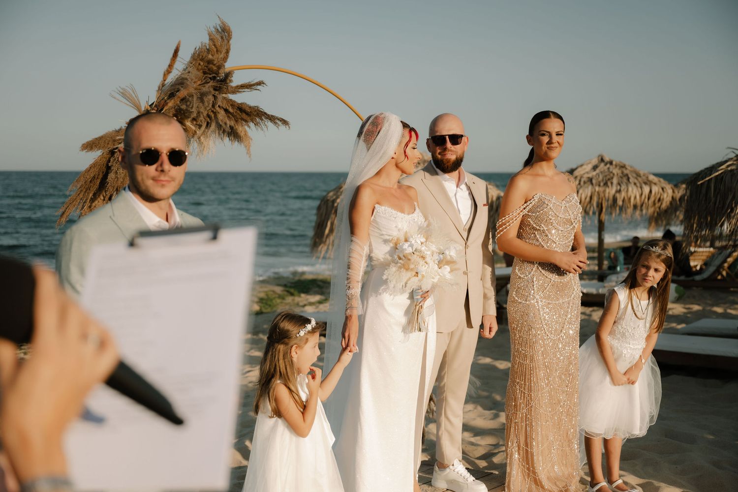 A beachside wedding celebration with people in neutral-toned attire gathered along the shoreline at sunset.