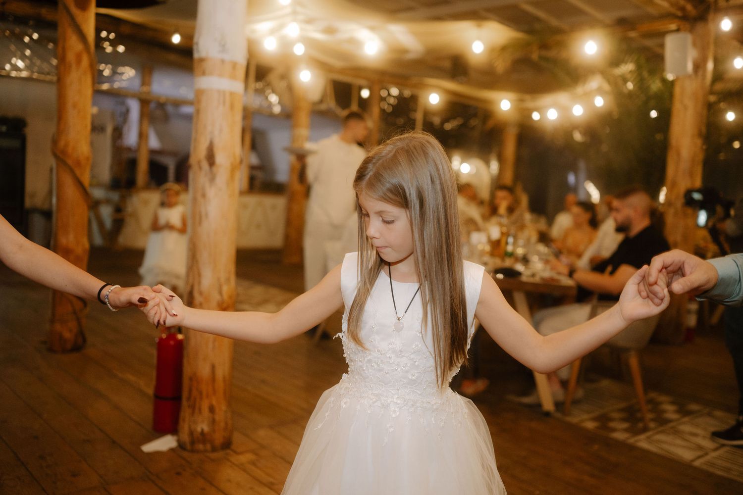 Wedding guests dancing and celebrating on a rustic wooden dance floor illuminated by warm string lights above.