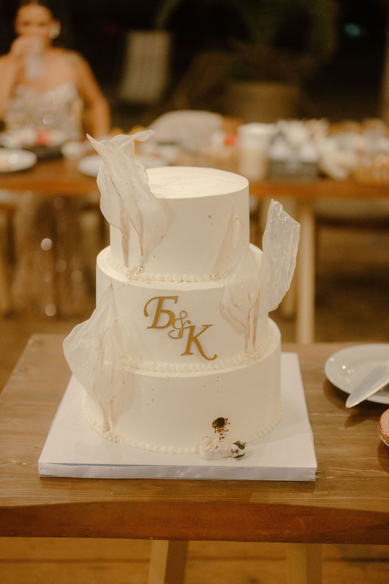 White wedding cake with elegant gold monogram initials displayed on wooden table at reception.