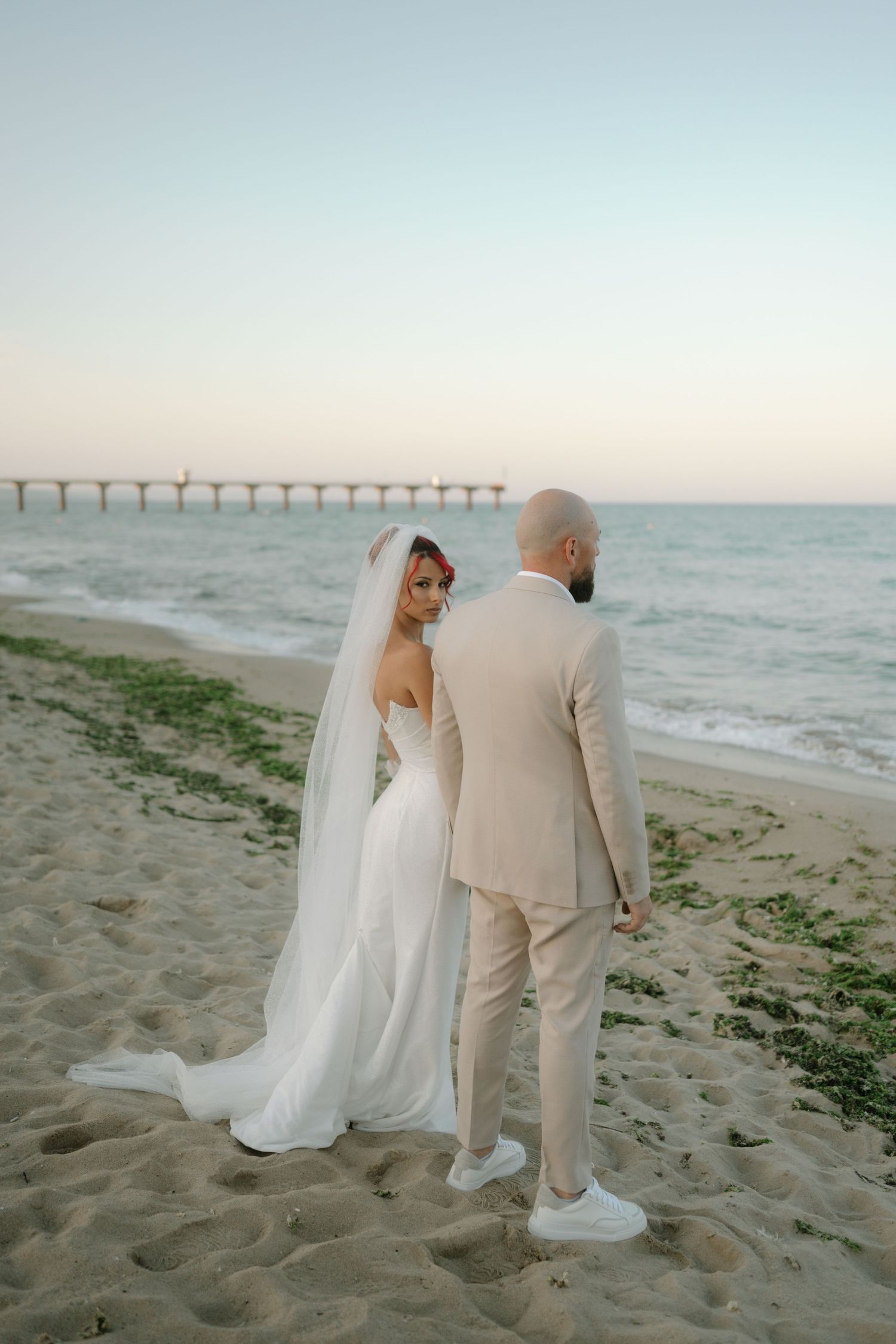 A couple in wedding attire stands on a sandy beach at sunset with a long pier visible in the distance.