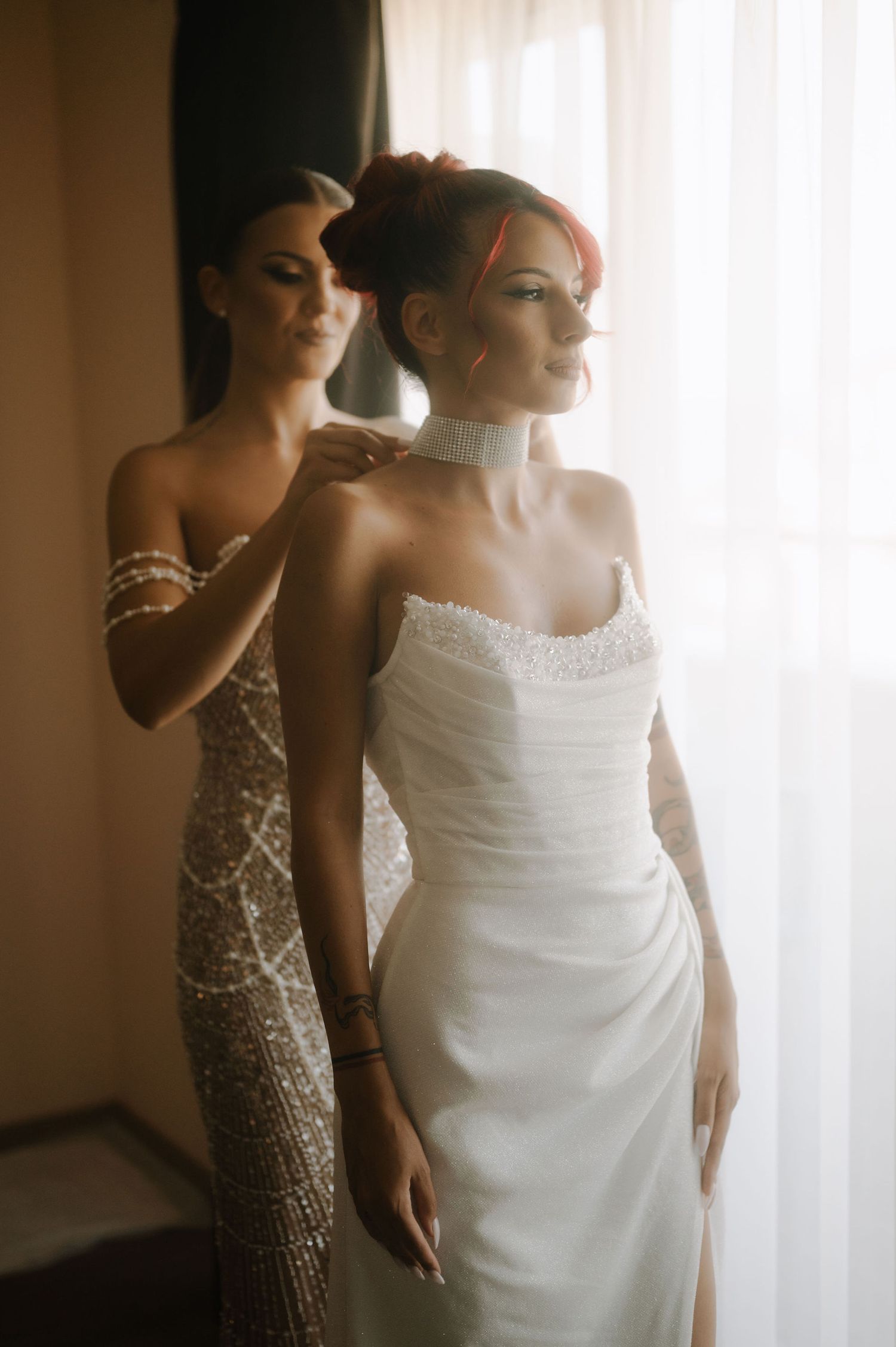 Bridesmaids in sparkly dresses help bride in white wedding gown prepare for ceremony in sunlit room.