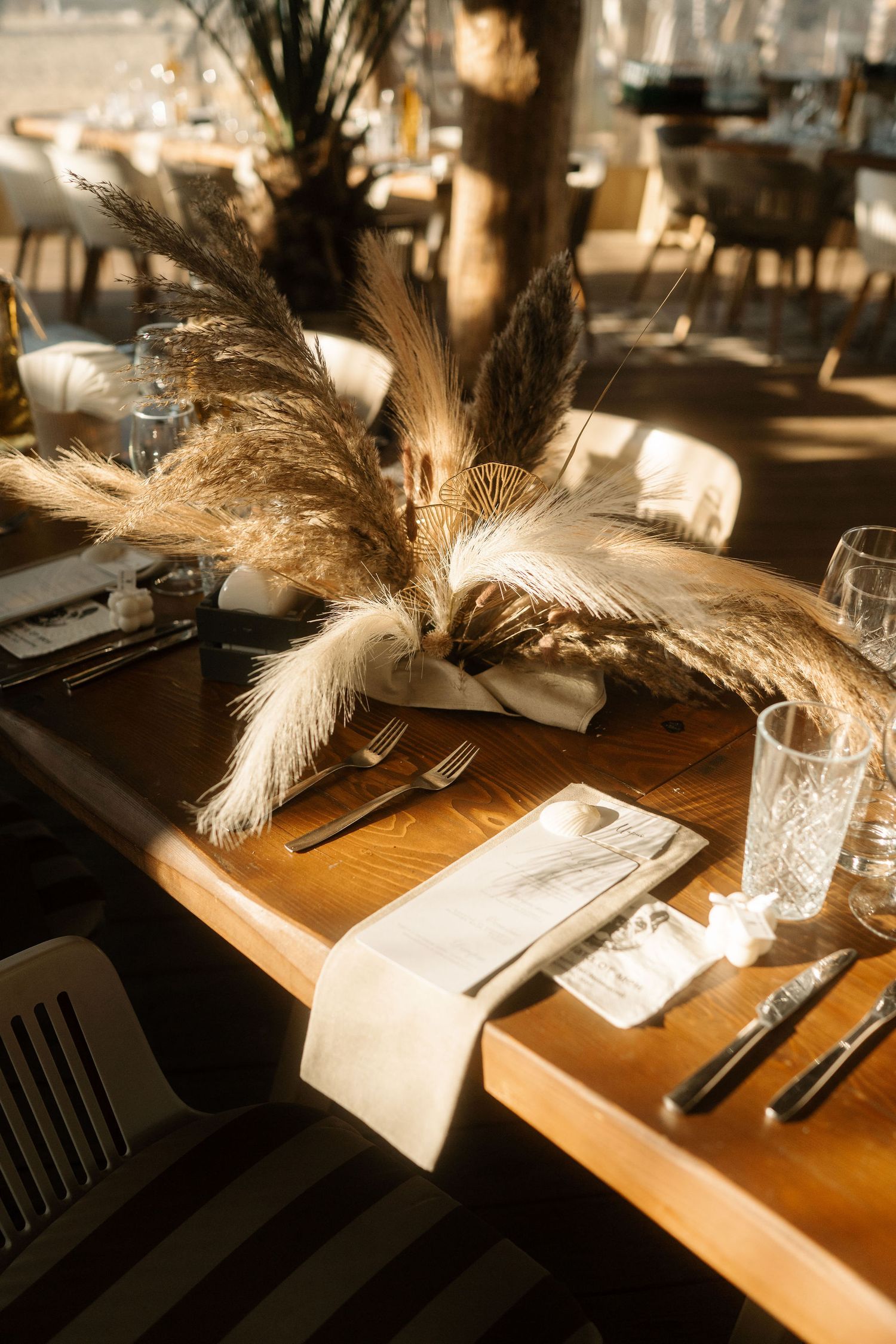 Elegant table setting with pampas grass centerpiece, silverware and glassware on wooden restaurant table in warm lighting.