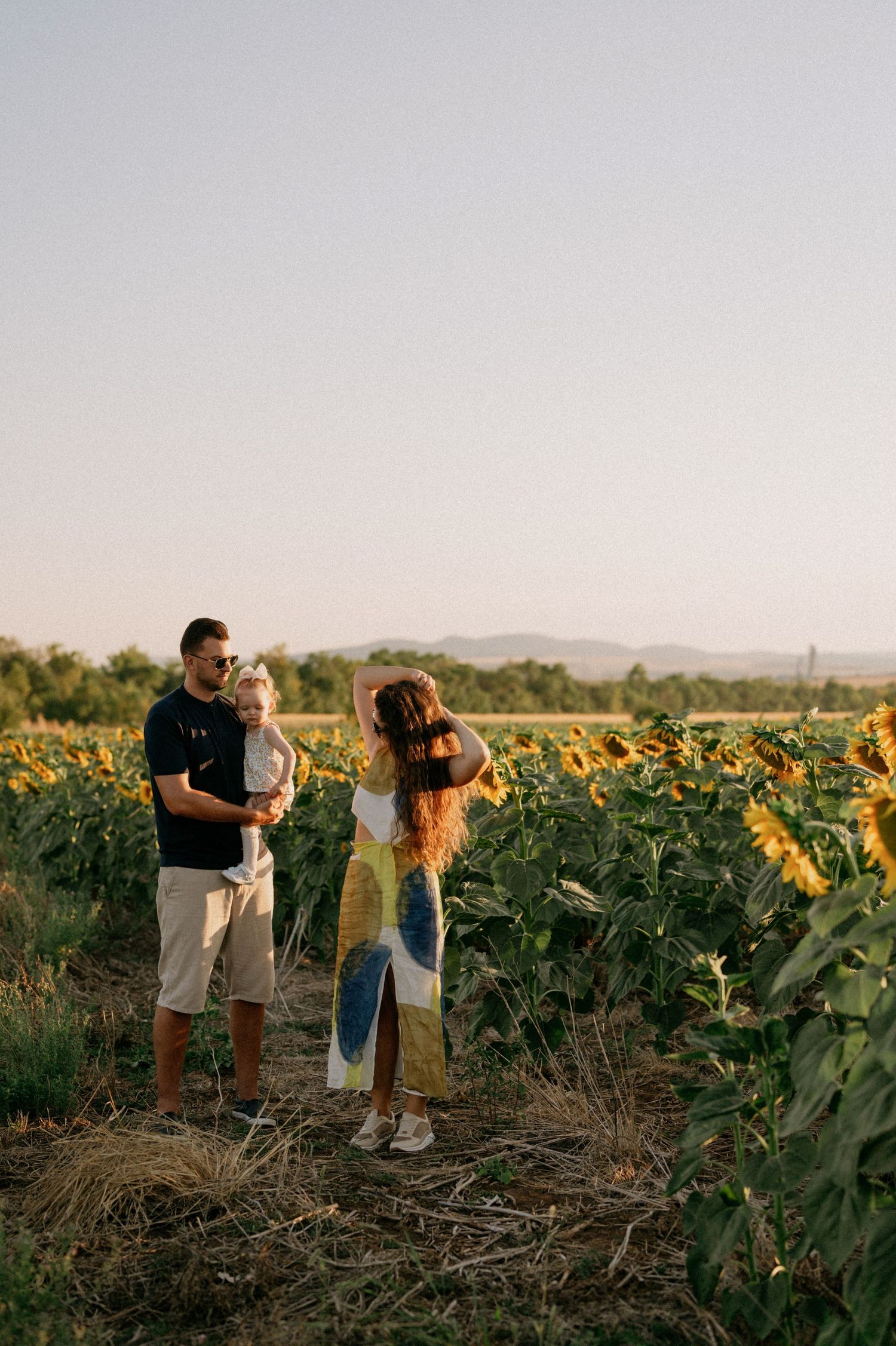Family of three standing in a sunflower field at golden hour, woman in floral dress, man holding baby.