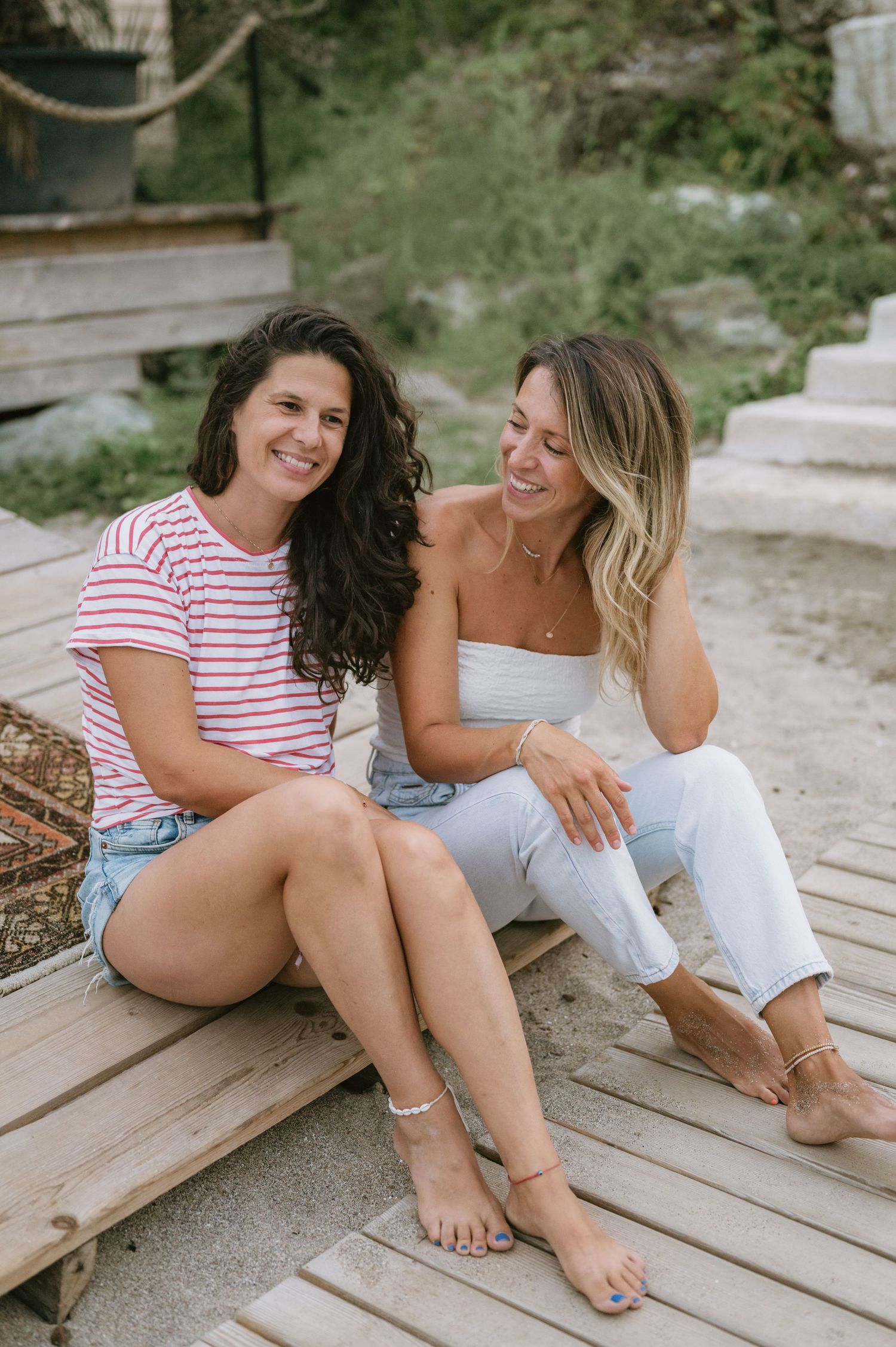 Two women smiling and sitting together on a wooden deck near a beach on a sunny day.
