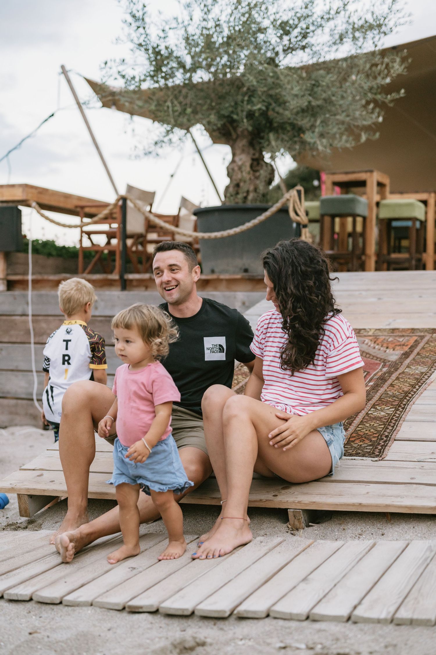 A happy family with two toddlers relaxing on a wooden deck near a glamping tent surrounded by olive trees.