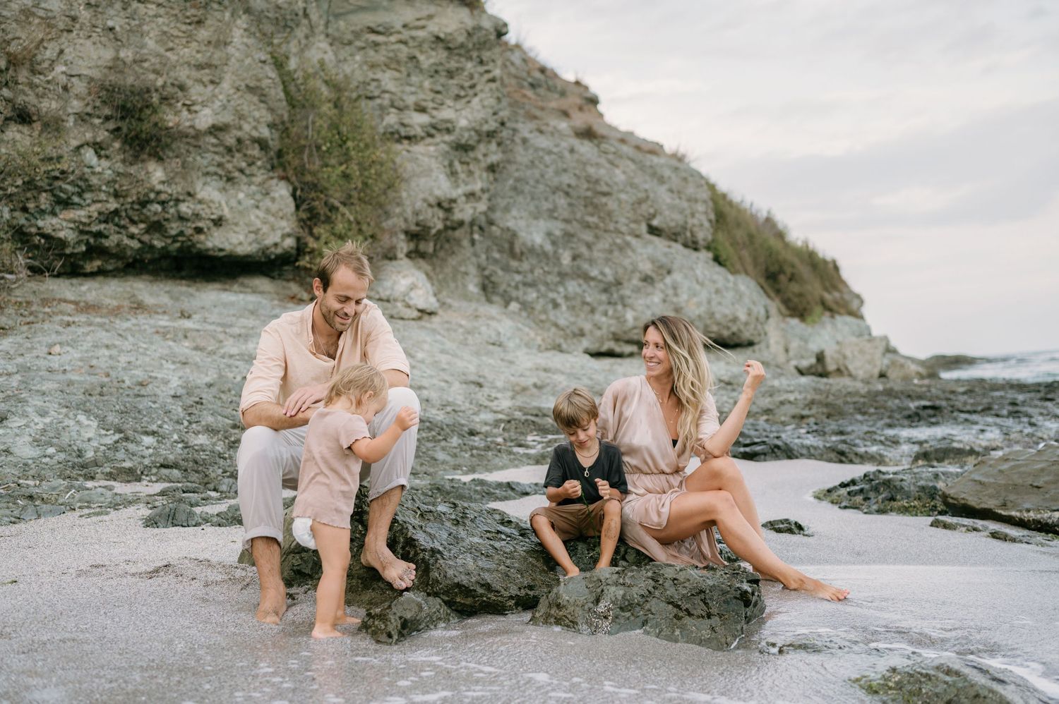 Family of four sitting on rocks at a sandy beach with cliffs, enjoying a relaxed outdoor moment together.