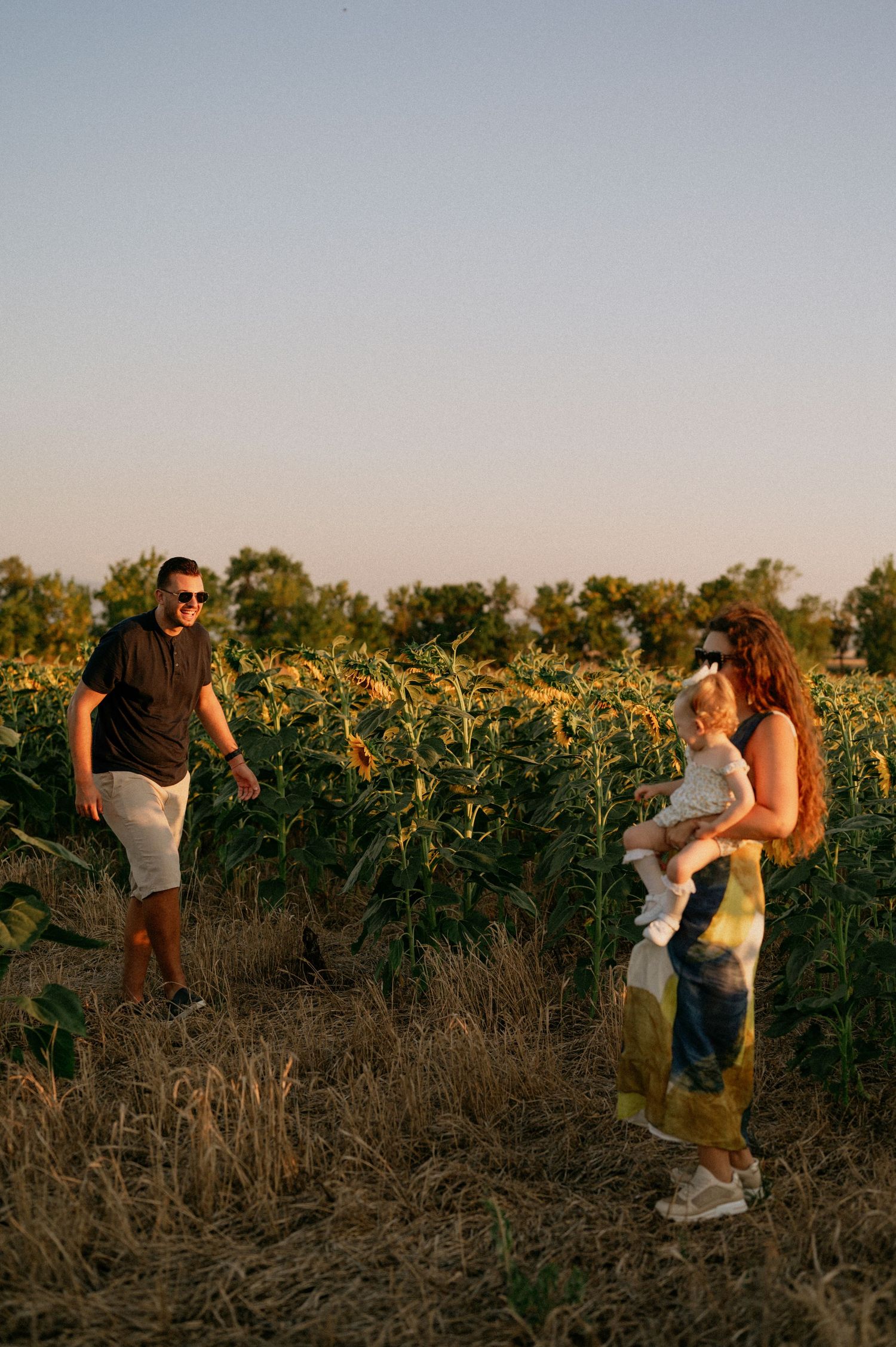 Family with baby enjoying golden hour in a sunflower field, parents smiling outdoors at sunset.
