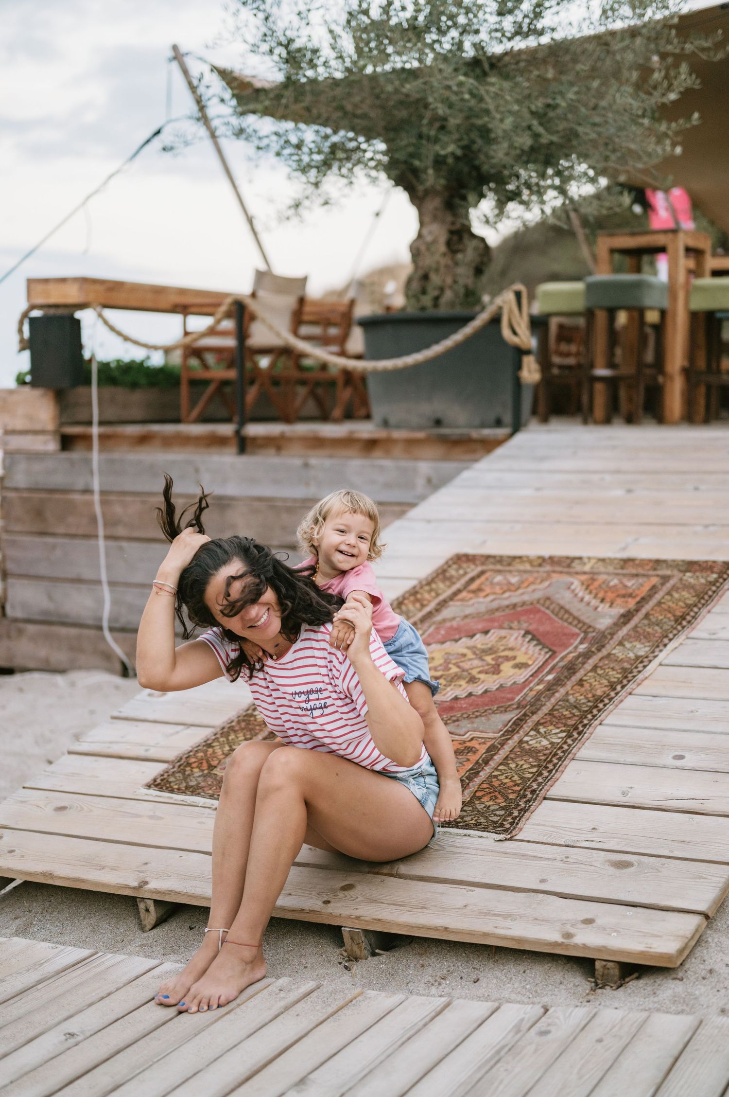 Woman sitting on wooden deck with cheerful toddler on her back, colorful rug and rustic outdoor seating area behind them.