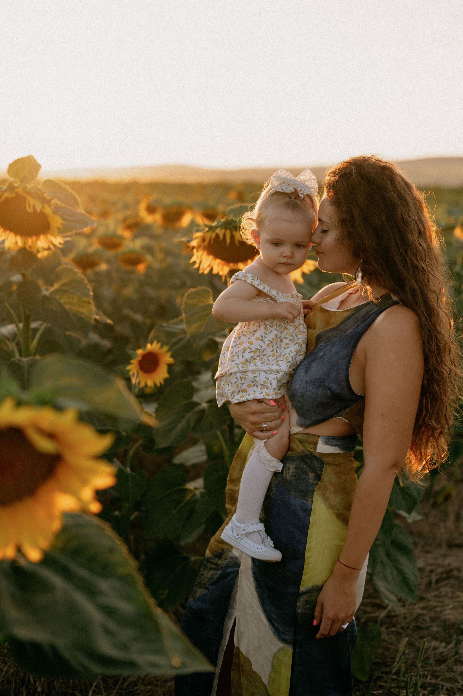 A mother holds her toddler in a golden sunflower field at sunset, gently kissing her cheek.