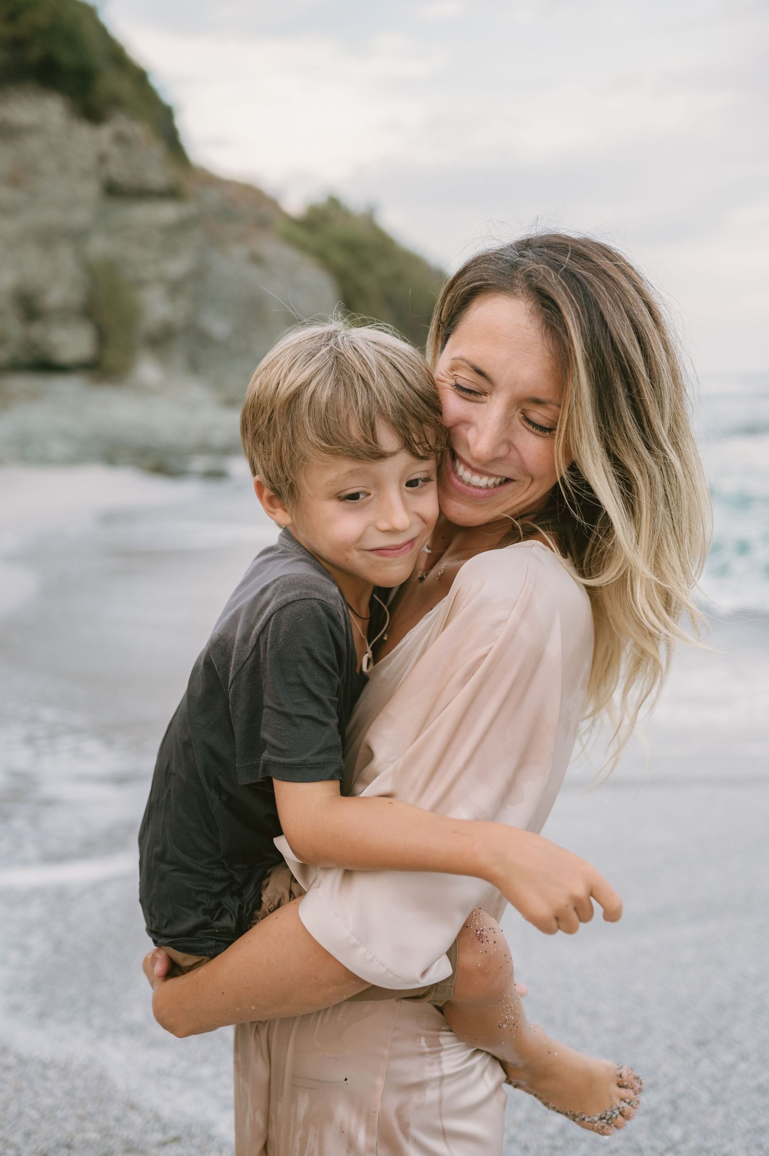 A smiling woman holds a young boy on a sandy beach with ocean waves and rocky cliffs in the background.