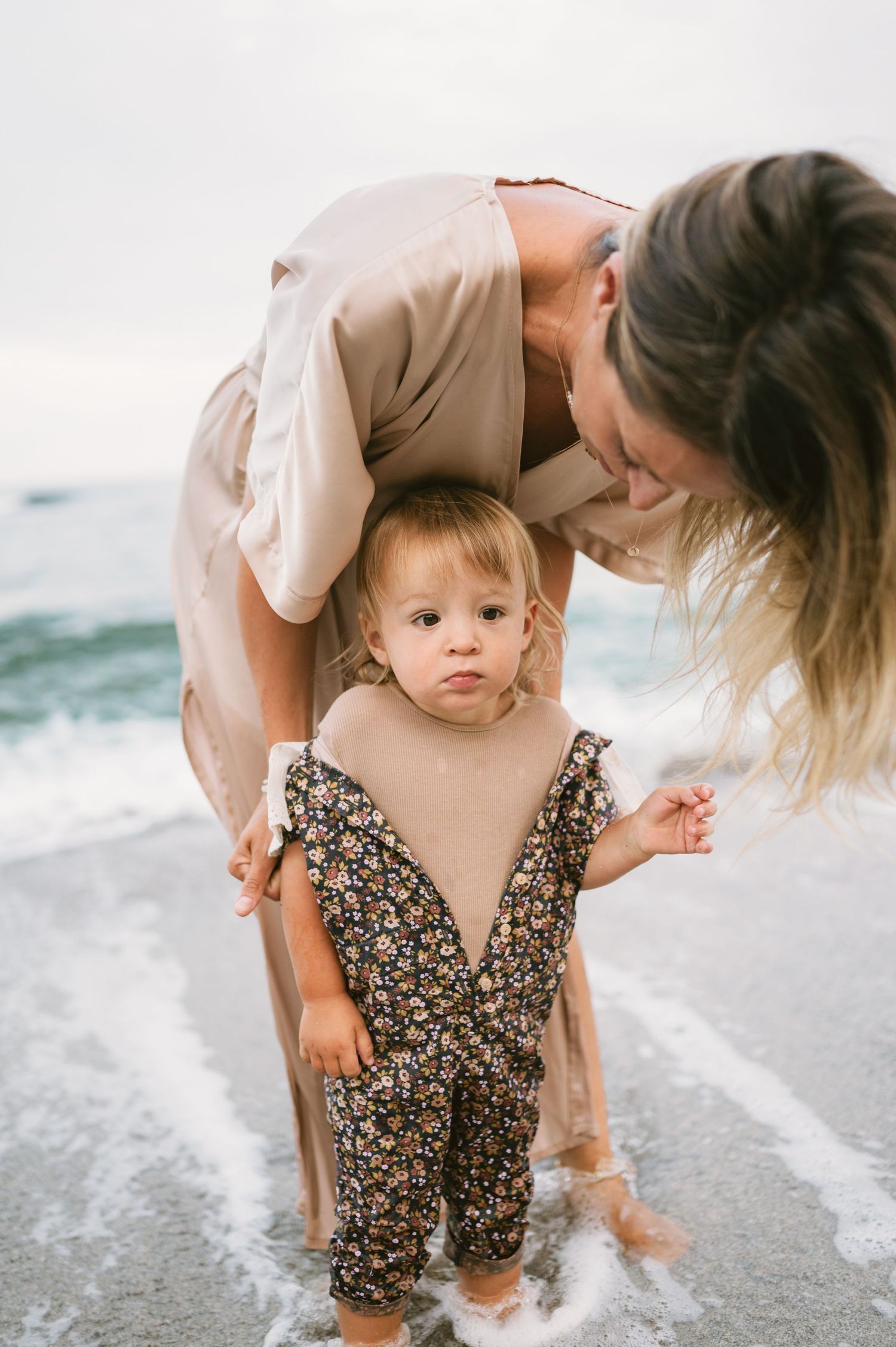 Mother helping toddler stand at ocean shoreline, baby wearing floral romper, waves washing over their feet at beach.