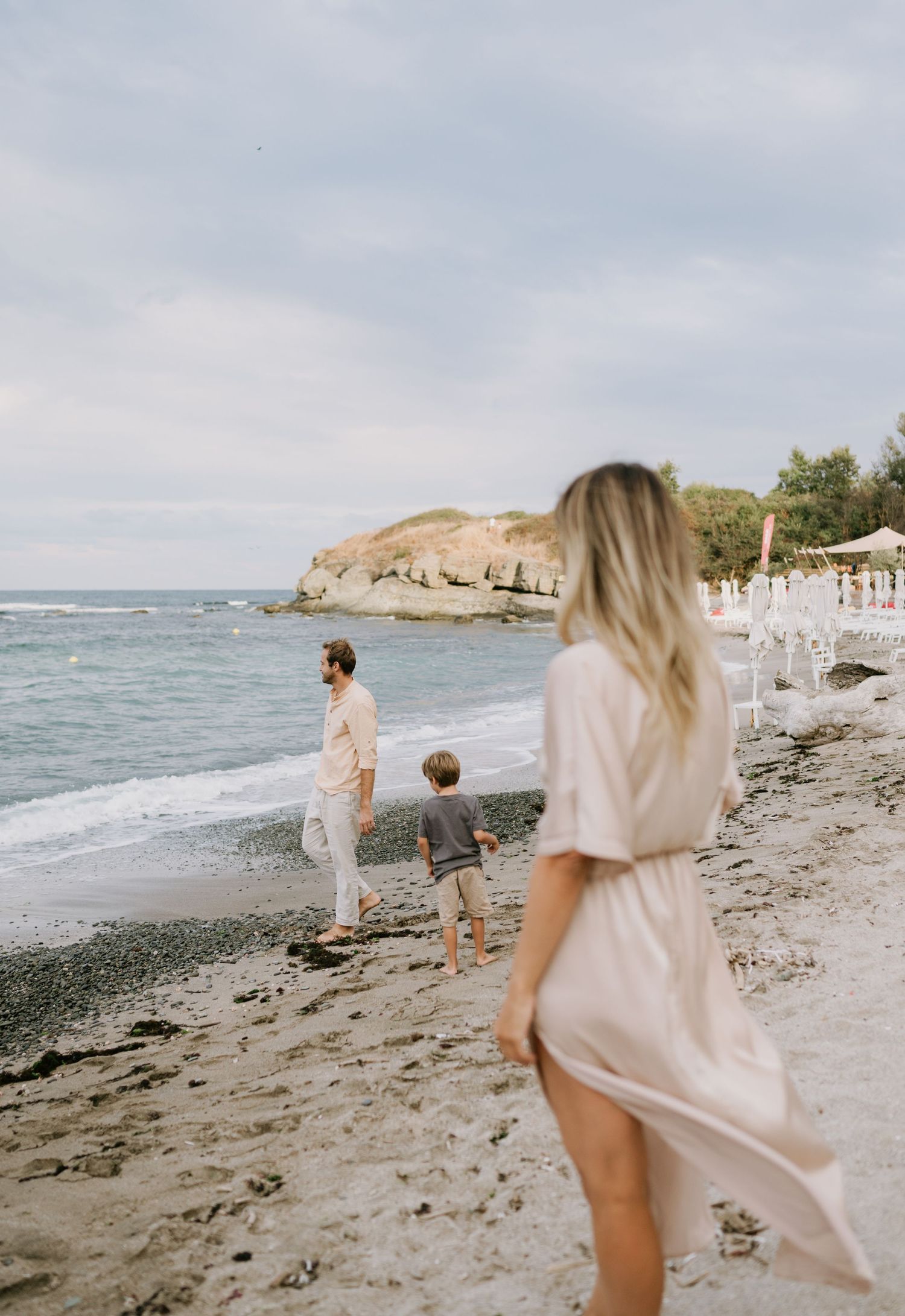 Family walking on a sandy beach near the ocean, with a rocky cliff and beach umbrellas in the background.