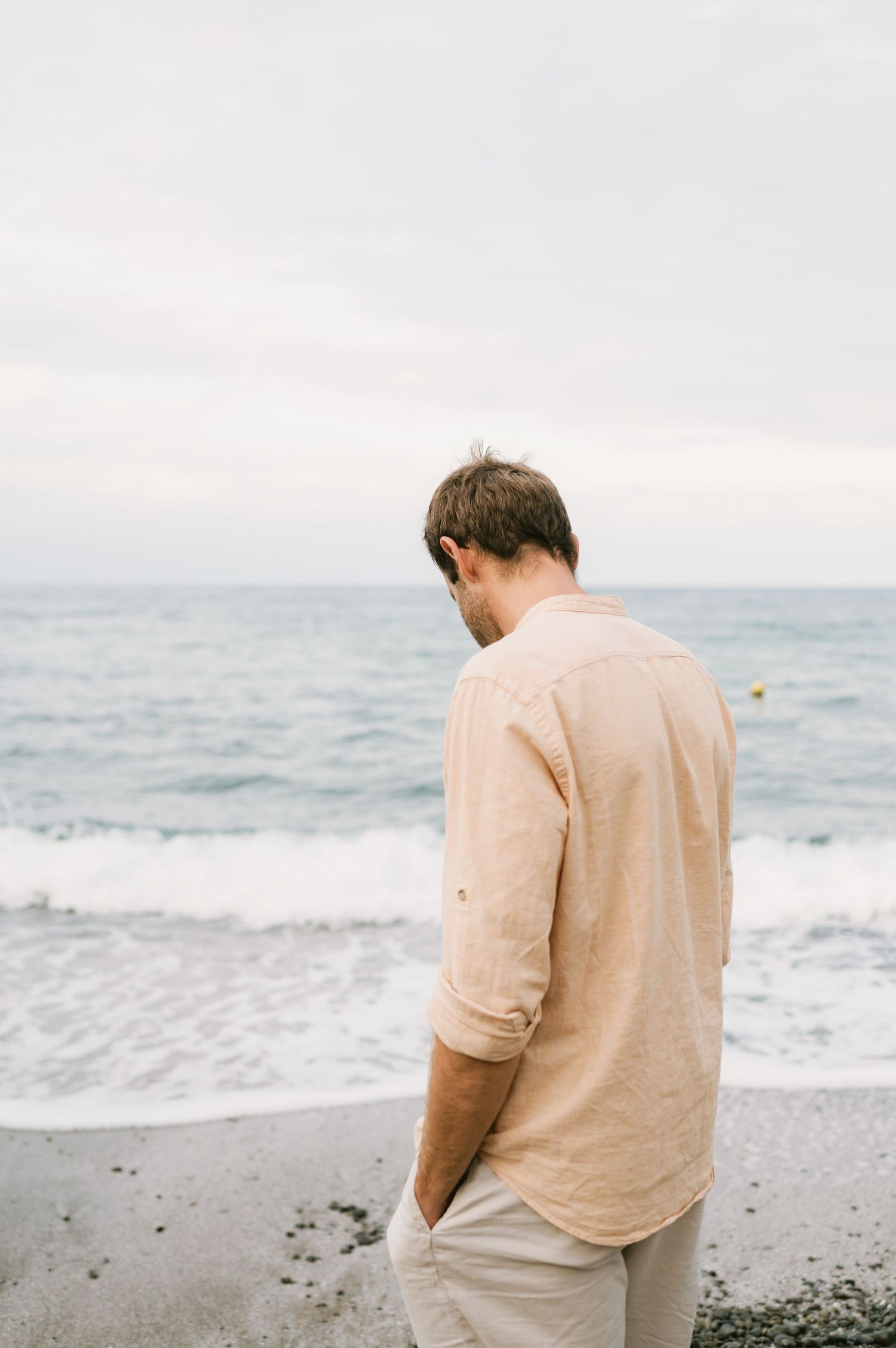 Man in beige linen shirt standing on rocky beach gazing down at shoreline with ocean waves and overcast sky behind him.