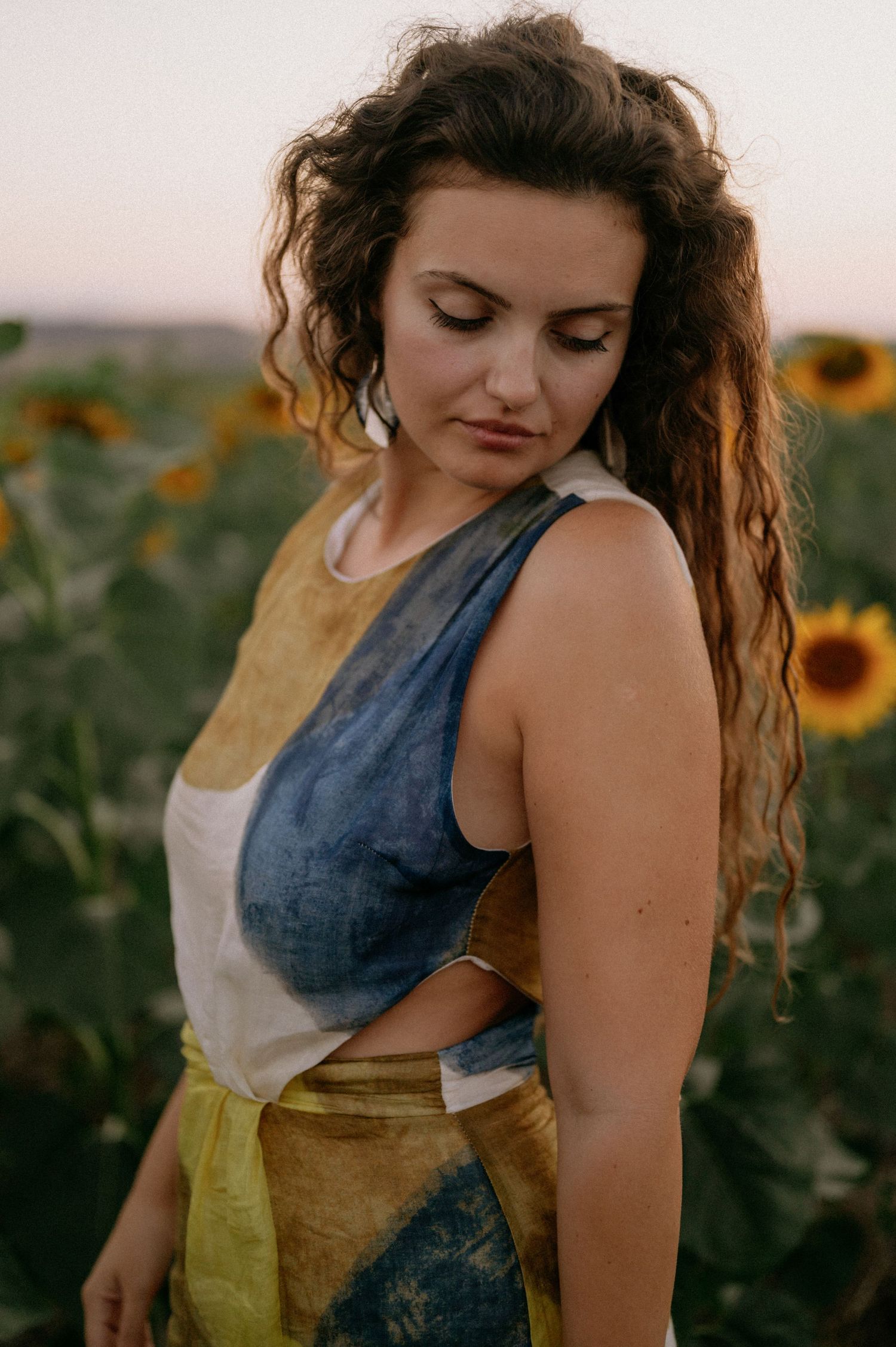 Woman in colorful patchwork dress standing in sunflower field at sunset.