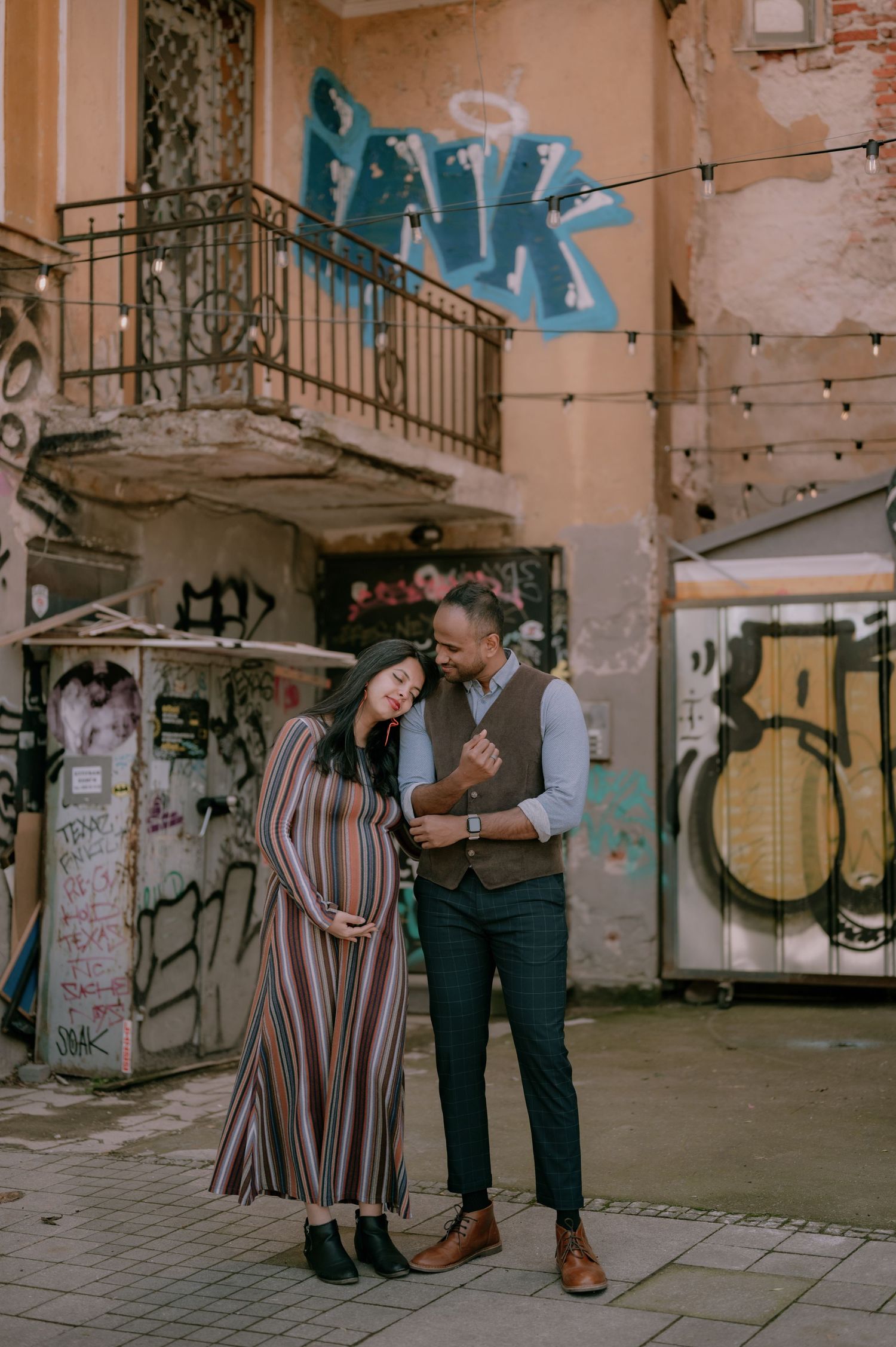 Pregnant couple poses tenderly in urban alley with colorful graffiti walls, woman in striped maxi dress, man in vest and trousers.